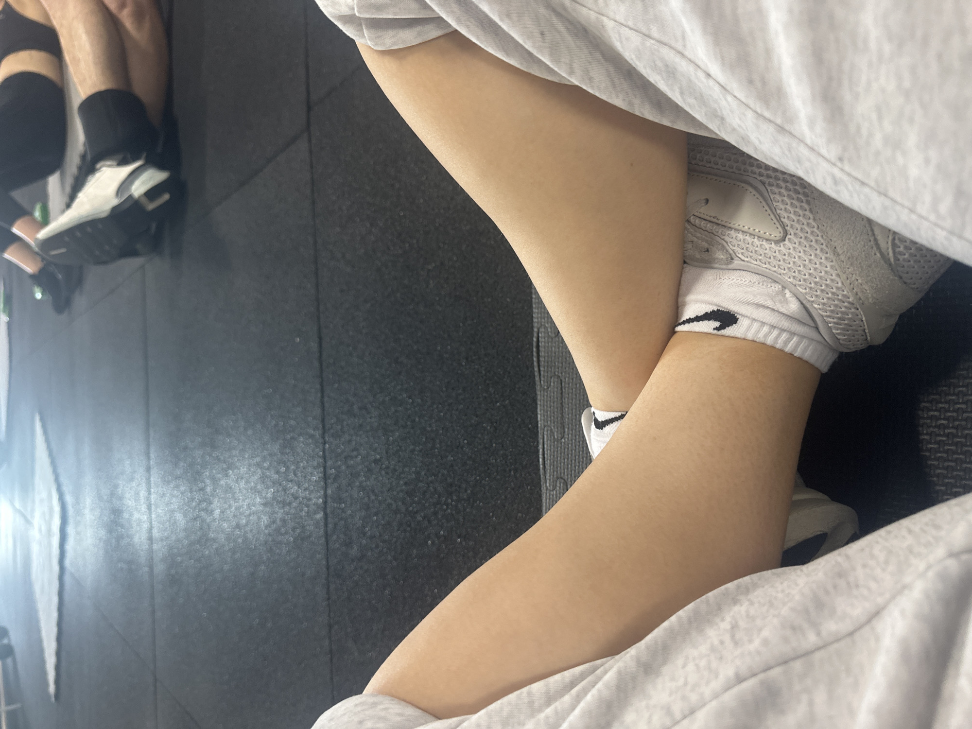The image shows a person sitting with legs crossed, wearing white socks with a black Nike swoosh and light-colored sneakers. The person is also wearing light-colored sweatpants or a similar garment. The background is a black, slightly reflective surface, possibly a rubber floor. In the upper left corner, part of another person's leg is visible, wearing a black sock and what appears to be a dark-colored brace or support. The location in Megion, Russia is unknown.