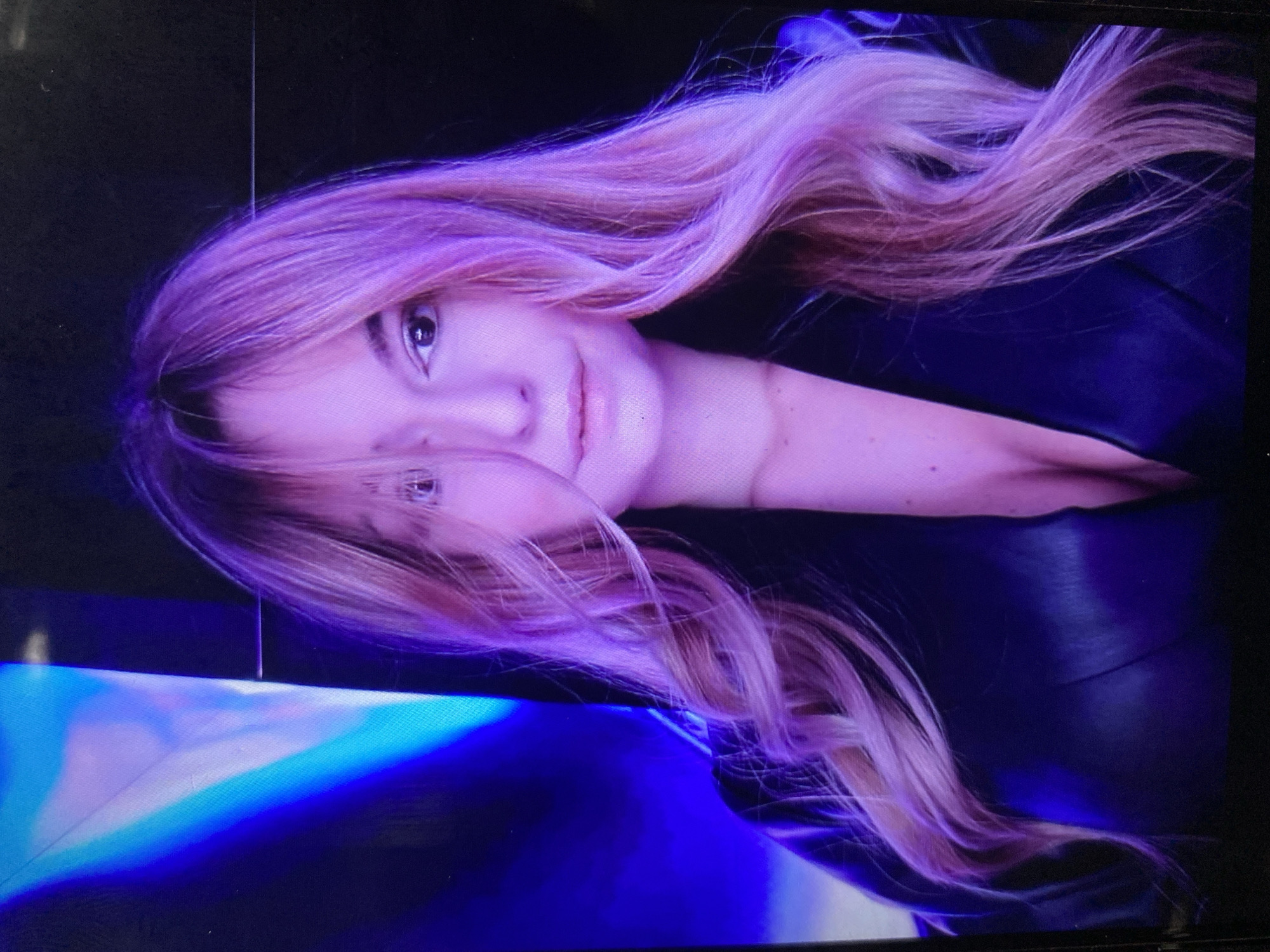 A headshot of a young woman is depicted, rotated 90 degrees clockwise, with her head on the left side of the frame. She has long, wavy hair colored in a vibrant purple or pinkish-purple shade, cascading around her face and shoulders. Her fair skin is illuminated with prominent purple and pink lighting, enhancing the color of her hair and creating an artistic glow.
The woman looks directly at the viewer with light-colored eyes, appearing to be brown or hazel, and sports a subtle, closed-mouth smile. She is dressed in a dark garment, likely a black top or jacket, with visible fabric texture and folds.
The background is mostly dark, suggesting an indoor or studio setting. A striking element is the bright, glowing blue and white light source positioned in what would be the bottom left corner if the image were correctly oriented. This light appears to be from a screen or stage effect, casting vibrant reflections. A thin, vertical line is visible in the dark background above the woman's head, possibly an artifact from photographing a digital display. The overall mood is stylized and theatrical, with the subject appearing calm and composed. No discernible text is present.