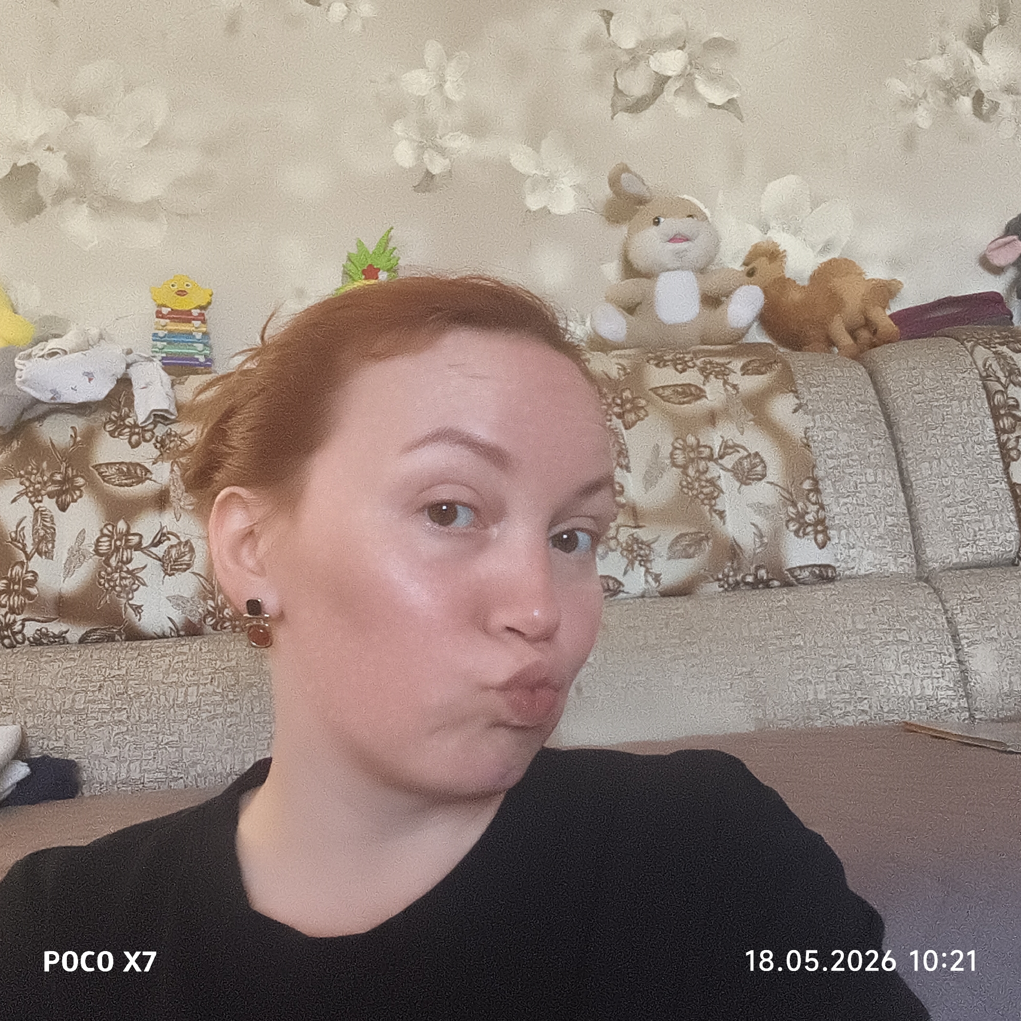 A selfie of a woman with red hair is taken indoors against the backdrop of a couch adorned with a floral pattern, upon which sit several stuffed animals including a bunny and a camel. To the left of the woman's head, there is a stack of colorful toys and baby clothes. The wall behind the couch is covered in wallpaper with a white floral design. The woman is wearing a black shirt and dark-colored earrings, and she is pursing her lips for the selfie. The bottom left of the image displays "POCO X7," while the bottom right reads "18.05.2026 10:21." No location details for Chaikovsky, Russia can be confirmed from the image.