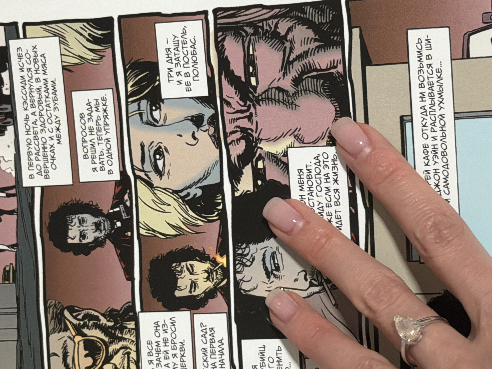 The image is a close-up shot of a page from a comic book. The comic panels feature illustrations of several male characters with dark, curly hair and beards, and one female character with blonde hair. The dialogue bubbles are in Russian. One panel shows a man with glasses and a beard, another shows a man smoking a cigarette with a flame. Another panel depicts a woman with her eyes partially closed. The bottom panel features a close-up of a man's face with a grim expression, and text in Russian. A woman's hand with light pink nail polish and a ring on her index finger is covering a portion of the comic page. The overall color palette is muted with brown and pink tones. The background appears to be a plain, light-colored surface. The image does not provide any information about the city of Moscow, Russia.