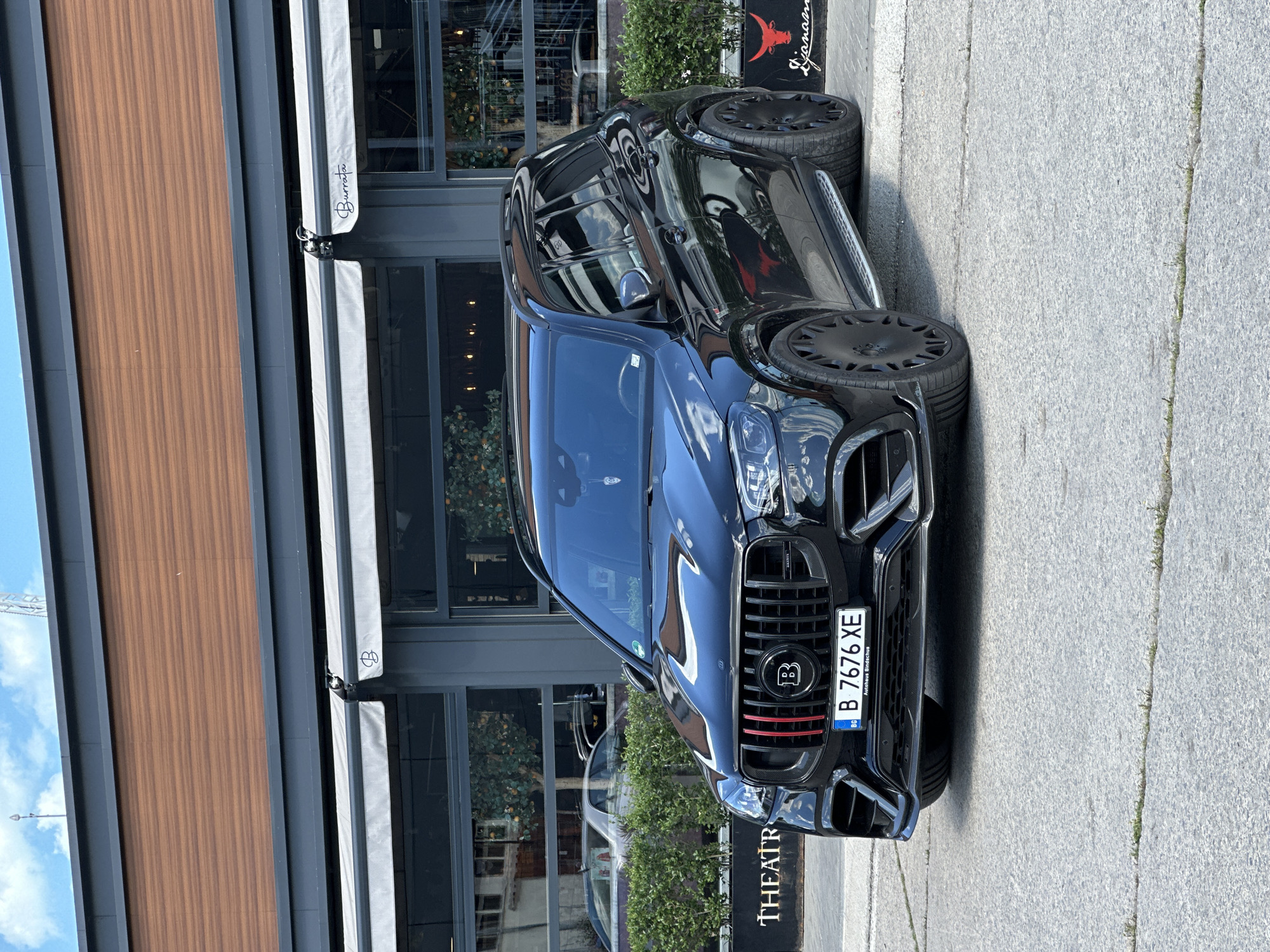 The image shows a black Brabus SUV parked on a city street in Varna, Bulgaria. The vehicle's license plate reads "B 7676 XE". Behind the car is a building with a wooden facade and dark-framed windows, some covered with white awnings labeled "Burrata". A sign on the ground near the car reads "THEATR". Another sign is visible with an illustration of a bull. The pavement is grey and shows signs of wear. No people are visible in the immediate foreground. The background hints at a lively urban environment.