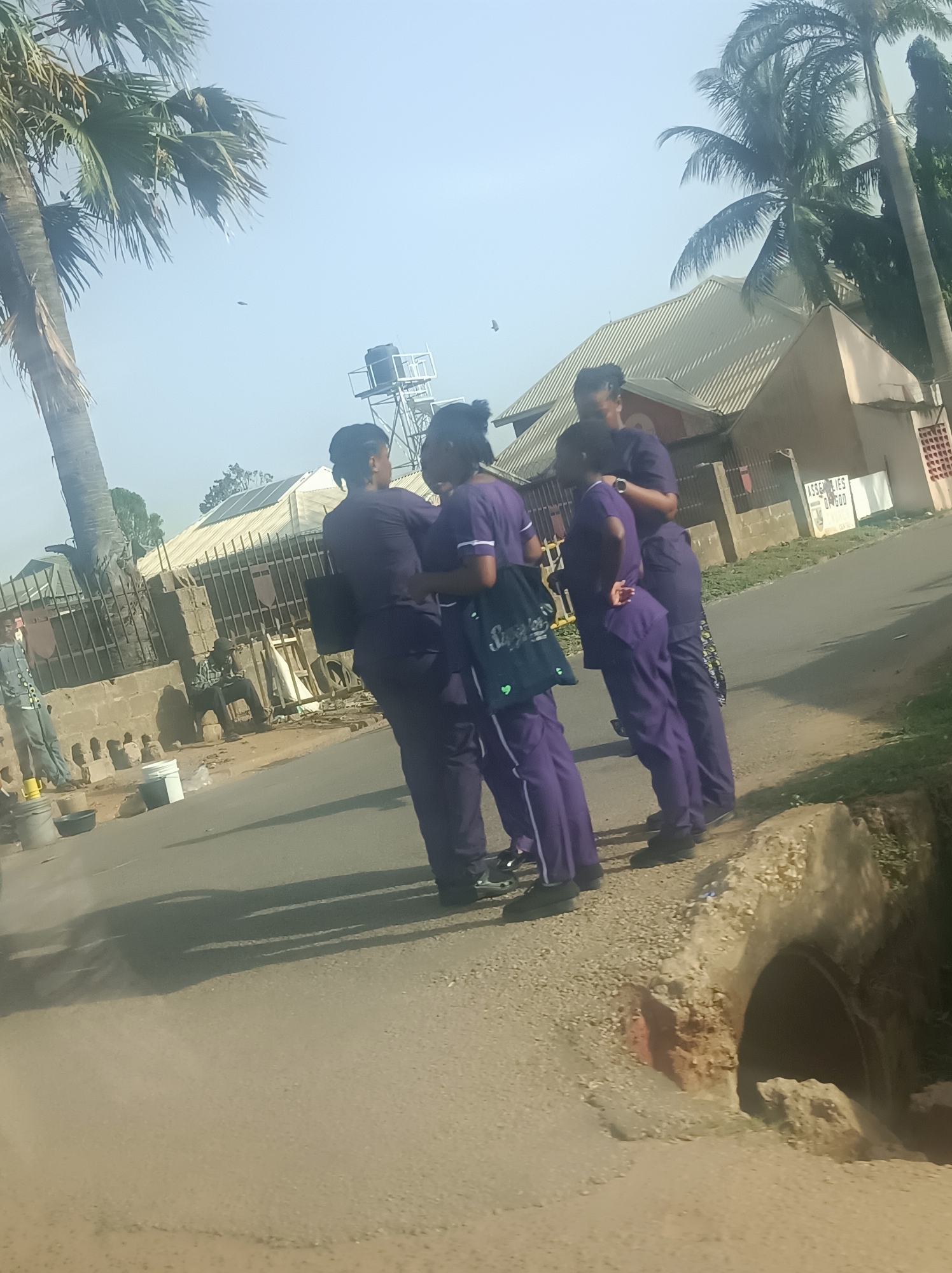 A group of four individuals wearing purple uniforms stand on the side of a road. A man is seated next to a metal fence and another person is standing next to him. Palm trees are visible in the background. A building with the words "ASSEMBLIES OF GOD" is also in the background. The location is in an urban area.
