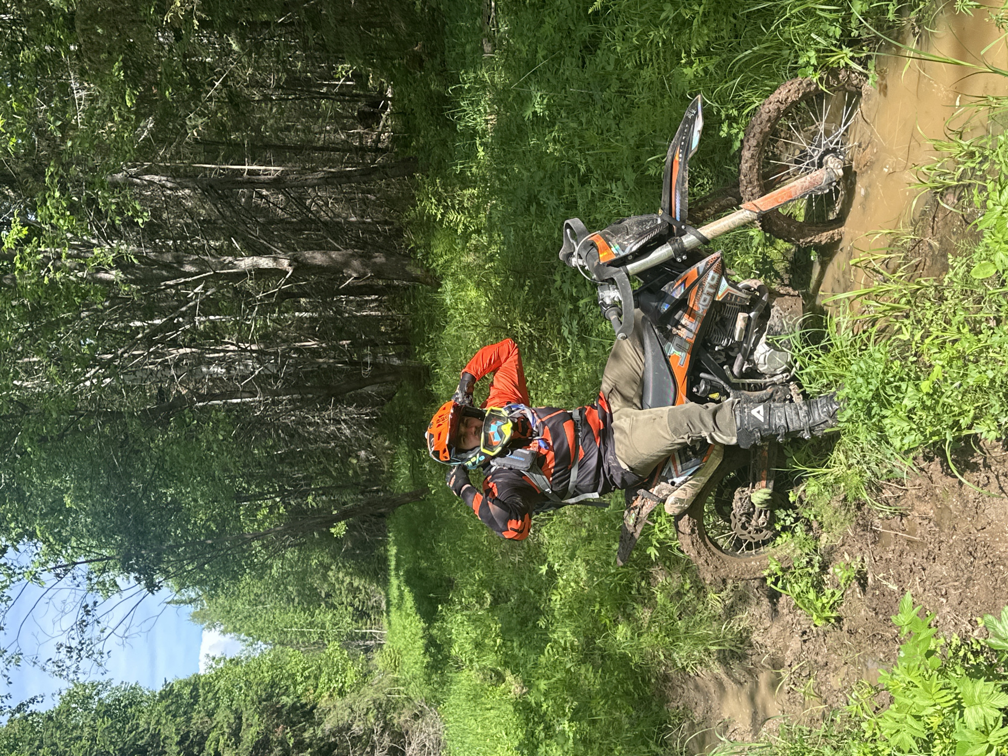 A motorcycle rider is stuck in a mud puddle on a dirt path in a forest near Ilyinsky, Russia. The motorcycle is leaning to its right side, with its front wheel submerged in the mud. The rider, wearing an orange helmet, goggles, and riding gear, is lying on the bike, turned towards the camera and making a gesture with his hands. The surrounding area is lush with green vegetation and trees. The path appears to be narrow and overgrown. The weather looks sunny.