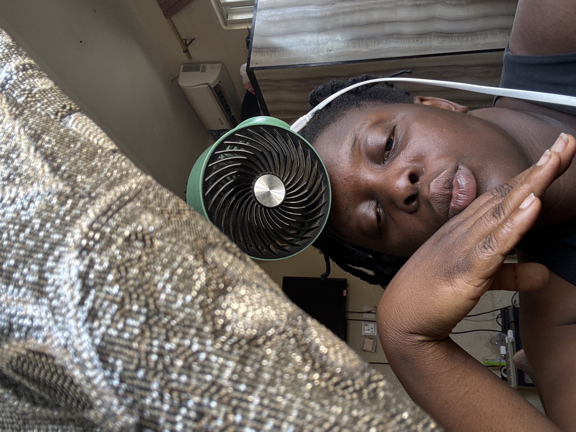 In Jawu, Nigeria, a person with dark skin and braided hair is captured in a close-up selfie. They are wearing a black tank top and have a small, green fan attached to their head. The fan is positioned on top of their head, with a white cable extending down and around their ear. Their face is turned slightly, with their lips pursed, and one hand is raised, touching their chin. In the background, a patterned fabric hangs from the top left, and other objects are visible, including an air conditioning unit, a cabinet, and a television screen. The scene appears to be indoors, with natural light entering from the right side of the frame.