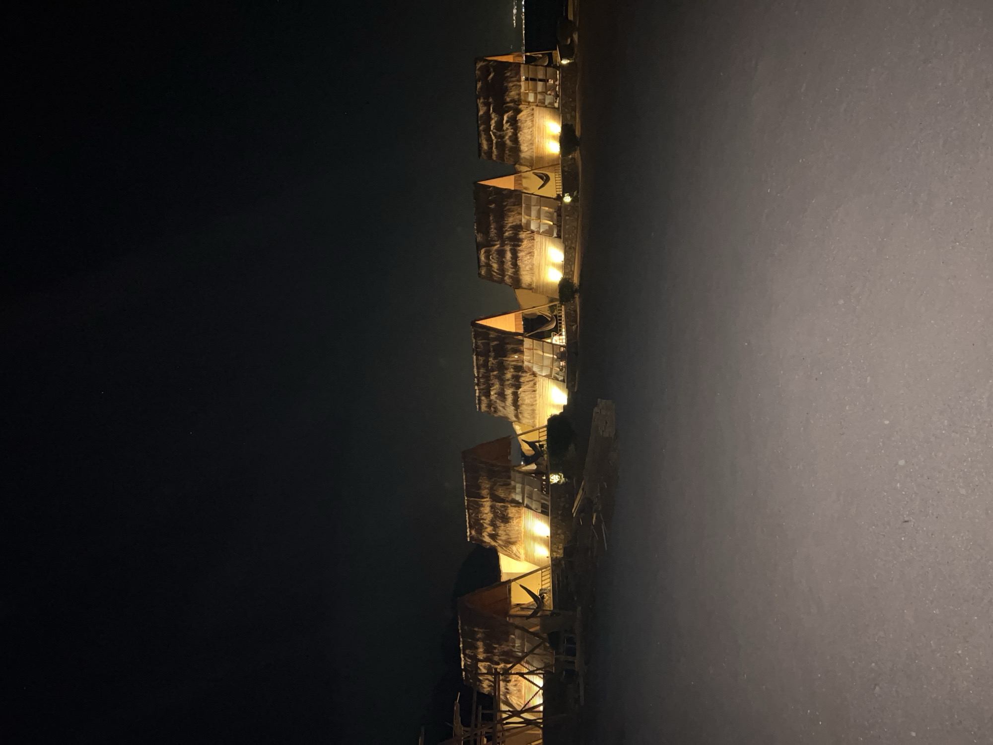 At night, a row of five A-frame huts with thatched roofs is illuminated by warm lights. The huts are situated on a sandy or dirt surface. Beyond the huts, a dark expanse of water is visible, with a faint hint of distant lights on the horizon, possibly indicating a shoreline or city. The sky is dark and clear, with a few faint stars visible. The scene appears to be in a resort or glamping area in Nueiba, Egypt.