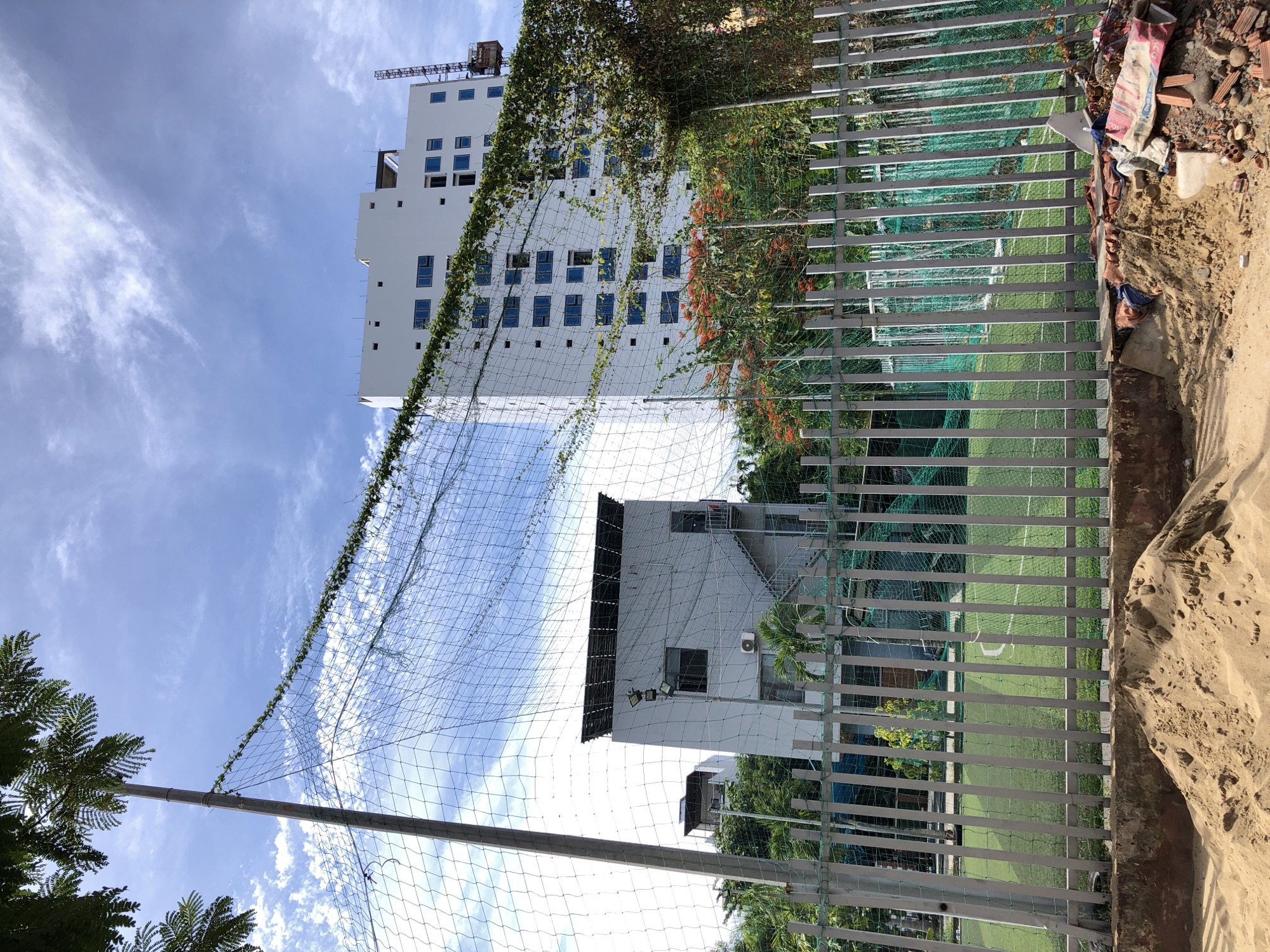 The image shows a fenced-off artificial turf sports field, possibly a small soccer field, in Da Nang, Vietnam. The field is enclosed by a tall metal fence and a green net. In the foreground, there is a pile of sand and construction debris. Behind the fence, two white buildings are visible. The building on the left is shorter and appears to be older, with a dark rooftop structure and an external staircase. The building on the right is a taller, modern apartment or office building under construction, with numerous windows. A construction crane is visible next to the taller building. Vegetation, including trees and vines, is present throughout the scene. The sky is blue with scattered white clouds. The scene appears to be an outdoor public space with elements of both recreation and ongoing development.