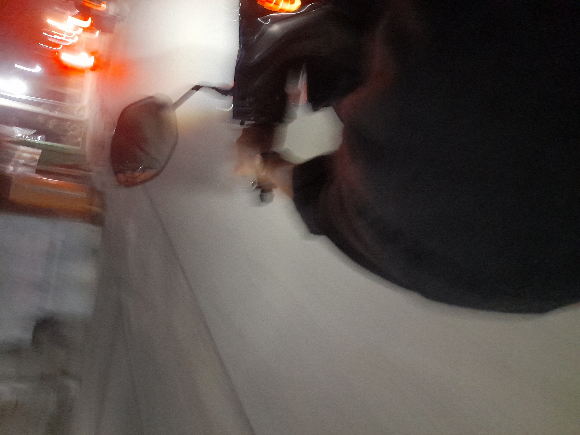 The image, captured at an unspecified time in Jetaksari, Indonesia, depicts a first-person perspective of someone riding a motorcycle at night. The foreground is dominated by the rider's dark jacket and a blurry view of their hands on the handlebars. To the right, a portion of the motorcycle's front, including a red taillight and a side mirror, is visible. The background is a streaky, motion-blurred scene of a street illuminated by bright lights, suggesting other vehicles or streetlights. The overall impression is one of movement and low-light conditions, typical of nighttime urban traffic.