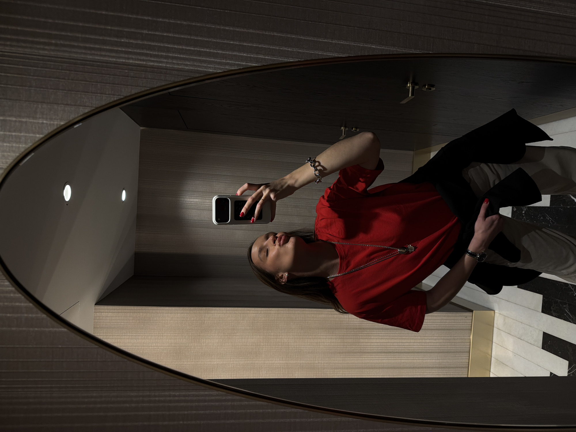 An indoor mirror selfie depicts a single individual, appearing female, in a hallway or room located in Budva, Montenegro. The subject has long brown hair and wears a red short-sleeved t-shirt, light-colored trousers, and a black jacket draped over her left arm. Visible accessories include a silver-colored necklace with a pendant, a silver-linked bracelet on her right wrist, a dark-faced wristwatch on her left wrist, and red nail polish on her fingernails. She holds a smartphone with a white case in her right hand, positioned to capture her reflection. Her head is tilted back, eyes closed, and lips pursed in a kissing gesture.
The environment, as reflected in the large, arched-top mirror, features walls covered in vertically striped textured wallpaper; one section is light beige, and another is darker grey. The floor displays a geometric pattern of black and white tiles. A gold-colored baseboard runs along the bottom of the wall. Two recessed spotlights are visible in the white ceiling. A dark wooden door with a gold-colored handle and lock mechanism is partially visible in the reflection.