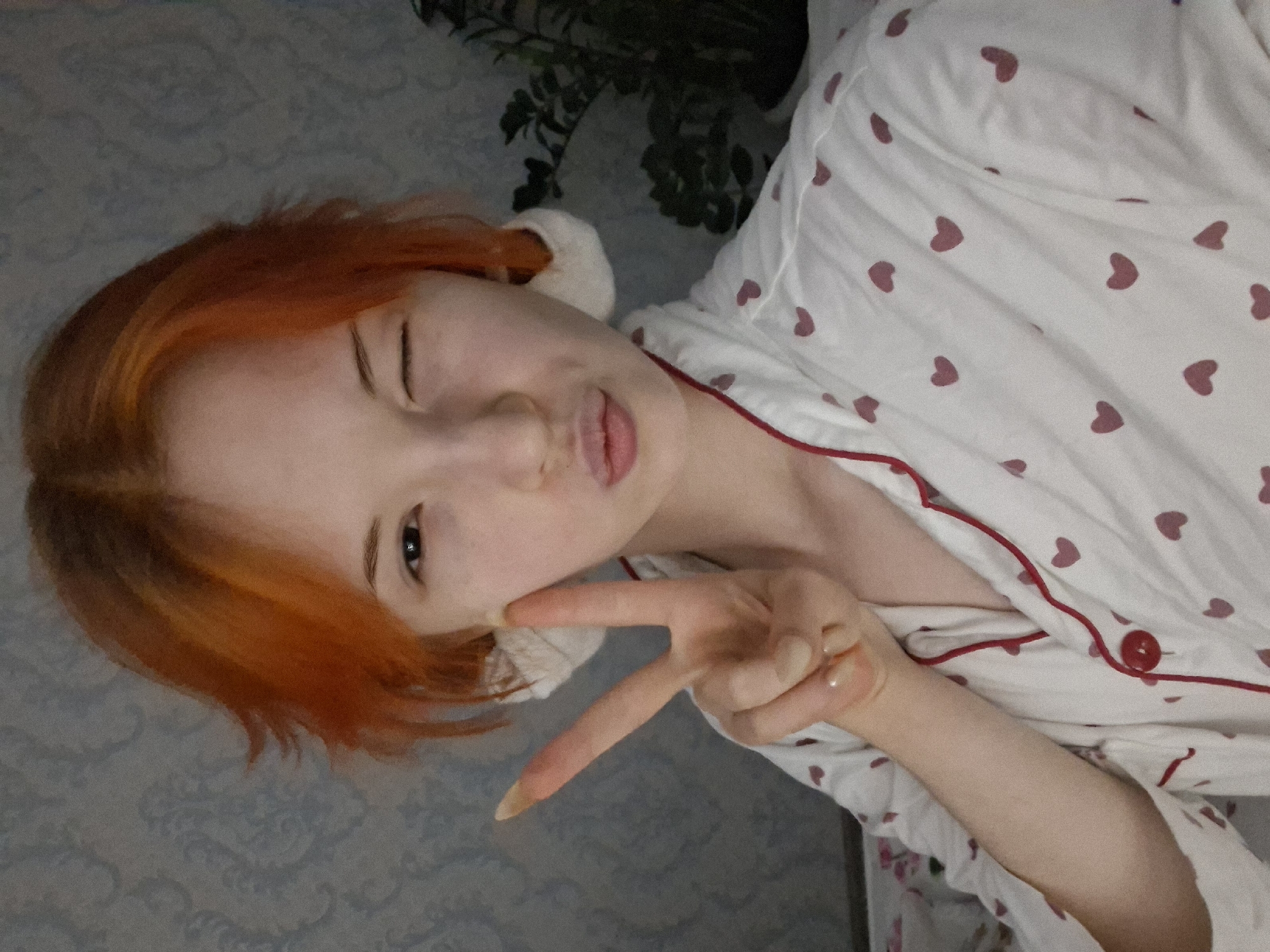 A young woman with short, orange hair is lying down, wearing white pajamas with red heart patterns and red trim. She is winking one eye and puckering her lips in a kissy face, while holding up a hand in a peace sign. The background includes a patterned wall and green plant foliage. There is no evidence to support that the location of the image is in the city of Chaikovsky, Russia.