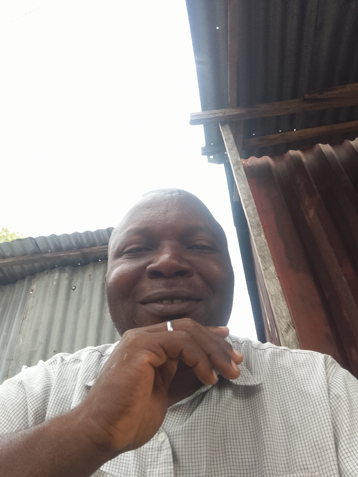 A medium-shot, high-angle selfie of a dark-skinned man with a shaved head, wearing a white, grid-patterned shirt, his hand up to his chin with a ring on his finger. Behind the man are corrugated metal sheets, some grey and some reddish, and some wooden support beams. The location is the city of Unknown, Unknown.