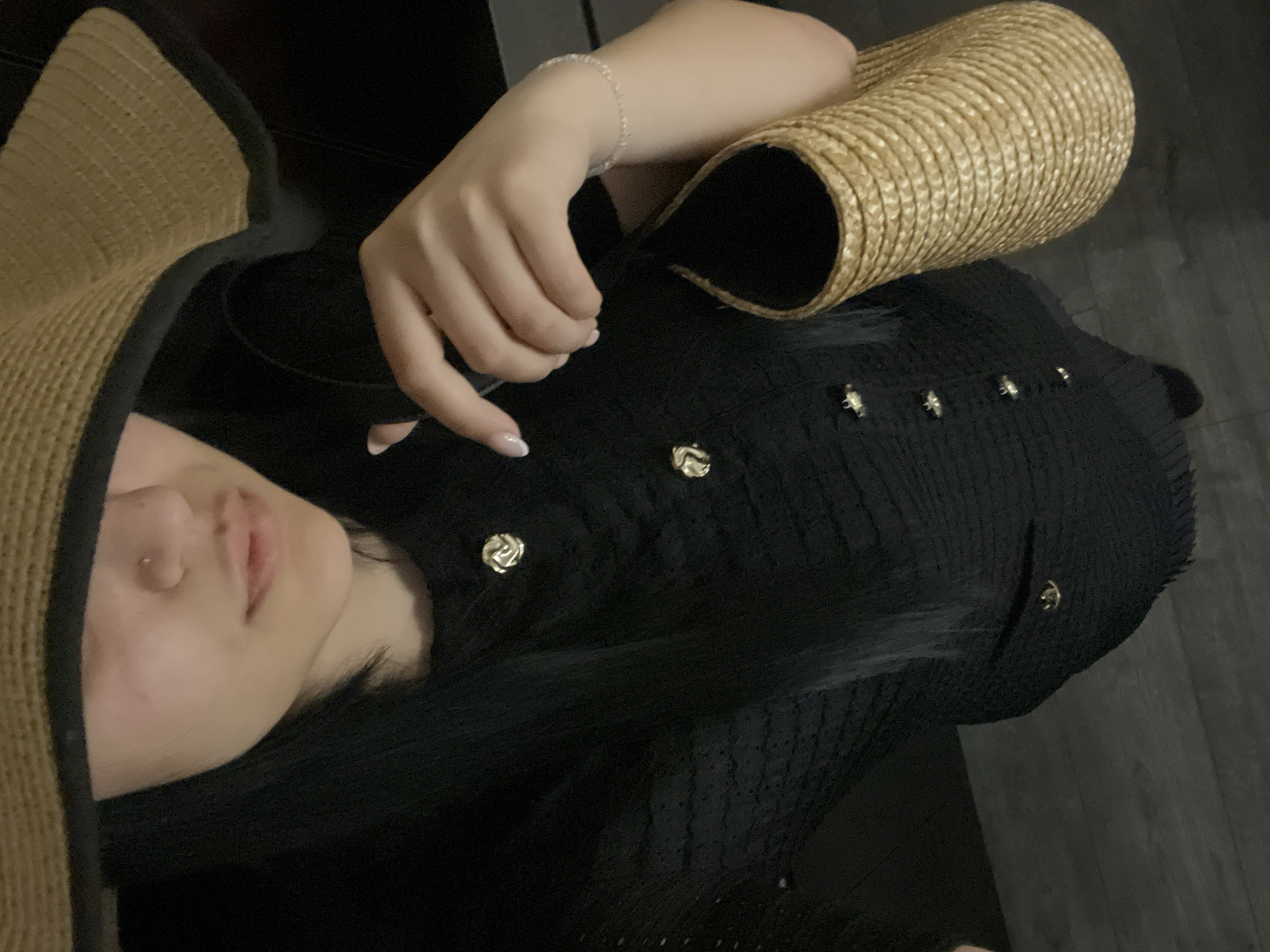This is a close-up, vertically oriented photo of a person lying down, possibly on a couch or bed, in an indoor setting. The primary subjects are the person's torso and upper body, a straw hat, and a black garment with decorative gold buttons. The person has fair skin and dark hair. They are wearing a delicate silver bracelet on their left wrist. The straw hat is held in their left hand and is positioned near their head. The black garment appears to be a jacket or top with a textured fabric and several prominent gold buttons, each designed with a floral or rose motif. The background is dark and indistinct, suggesting low lighting conditions, potentially indicating evening or nighttime. There are no visible text elements in the image. The overall mood is relaxed and intimate due to the close-up perspective and the subject's reclined pose. The specific location of Tvarditsa, Bulgaria, is noted, but no visual cues within the image itself directly support this context.