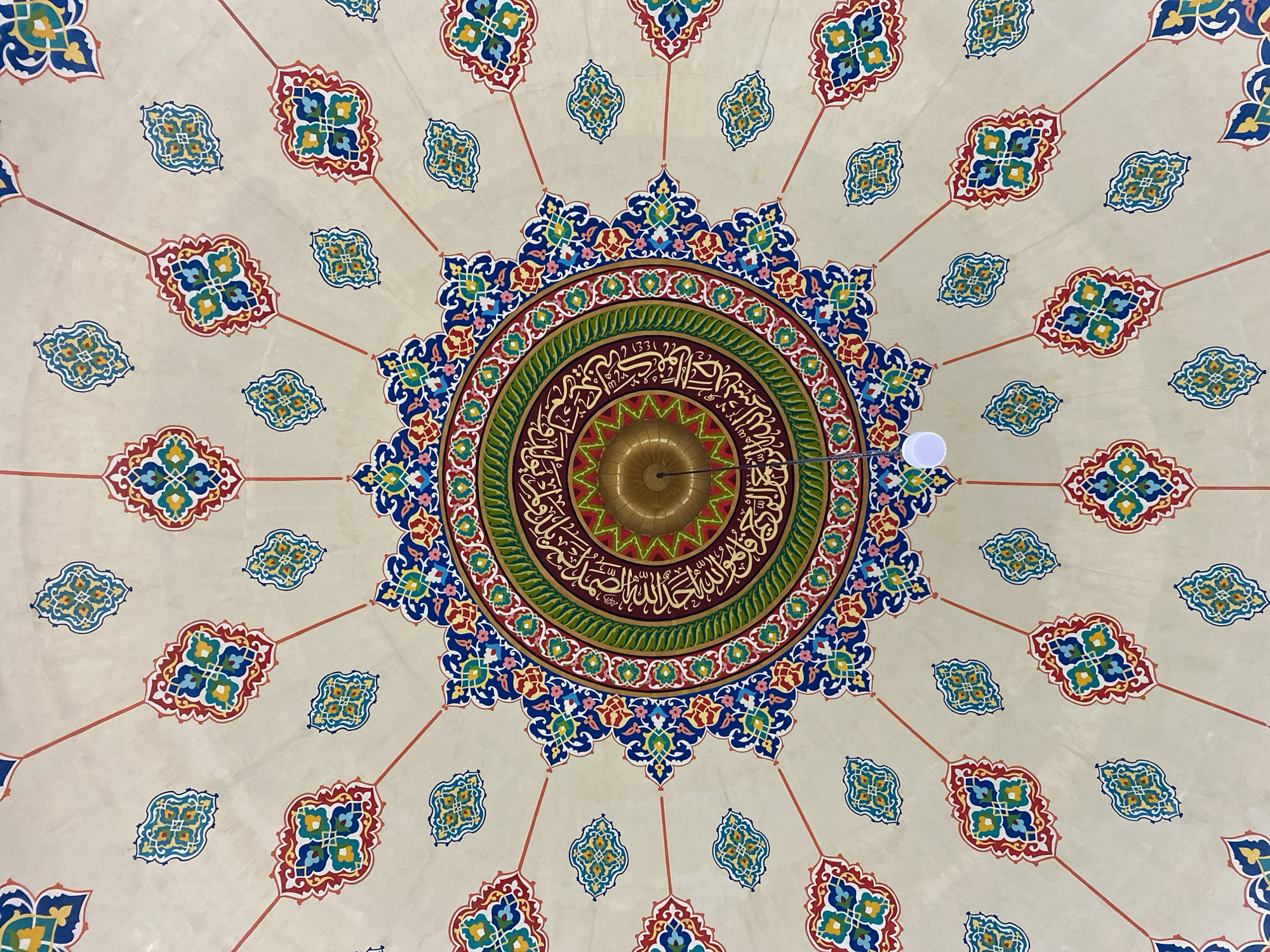 This image is a close-up, downward-facing shot of a highly decorated ceiling, likely in a mosque. The ceiling features a central, intricate circular motif with radiating lines that connect to smaller, repeating ornamental patterns spread across the light-colored surface. The central design is a complex mandala-like pattern in vibrant colors of red, blue, green, yellow, and gold, with Arabic calligraphy encircling a golden dome-shaped fixture. The smaller, repeating patterns are also colorful, featuring geometric shapes and floral elements. Two lights are visible; one is the golden fixture at the center, and another is a small white light hanging from a wire. The overall impression is one of ornate Islamic art and architecture. The image was taken at Masjid Agung Islamic Center Lhokseumawe in Lhokseumawe, Indonesia.