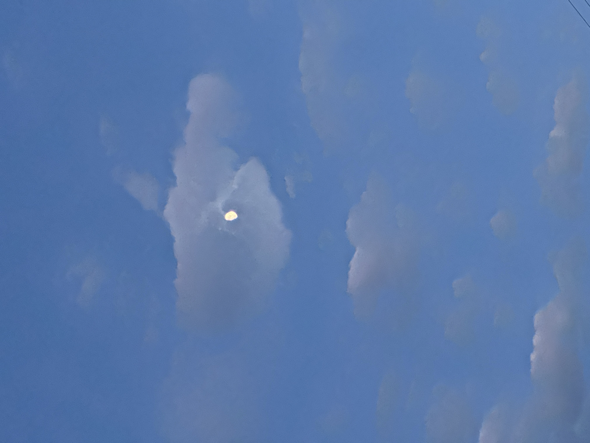 The image captures a daytime sky with scattered clouds. The sky is a clear blue color. Several fluffy, white and grey clouds are visible, with one notable cloud obscuring a portion of the sun, creating a halo effect. Thin, dark wires run along the top right of the photo. The location is unknown.