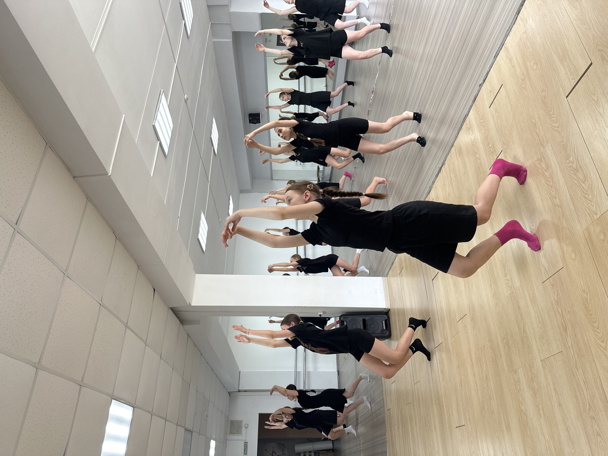 In a dance studio, located in Votkinsk, Russia, a group of young women in black athletic wear are practicing a choreographed dance. The studio features a light-colored wooden floor, large mirrored walls, and a white drop ceiling with fluorescent lighting panels. Several students are visible in the foreground and background, all engaged in synchronized movements. Their poses suggest a graceful, flowing dance routine. One dancer in the immediate foreground is captured mid-movement, with her arms extended upwards and her body bent. Another dancer to her left mirrors a similar pose. The reflection in the mirrors provides a sense of depth and expansiveness to the room, showing multiple angles of the dancers' practice. A black speaker is visible in the background near a pillar, and a "EXIT" sign is mounted on a wall.