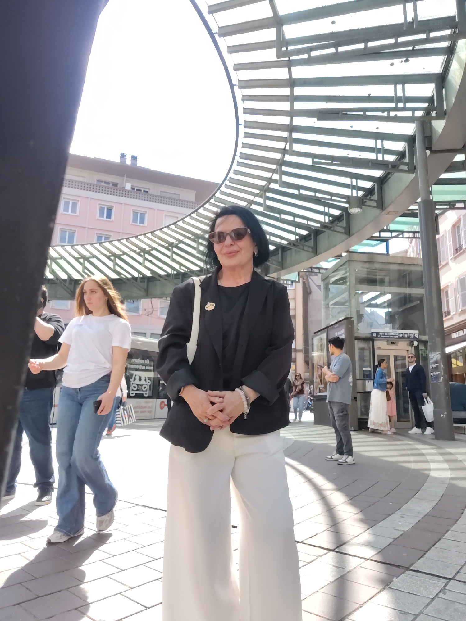 A daytime scene shows a woman in the foreground, standing outdoors on a paved public space marked with circular patterns. She wears sunglasses, a dark blazer, and white wide-leg pants. Other people walk in the background. Behind the woman is an open-air steel canopy structure. Across the street are building facades with windows and architectural details, indicative of a European urban environment. A glass elevator structure is visible. The image was taken in Place de l'Homme de Fer, a transportation hub in Strasbourg, France.