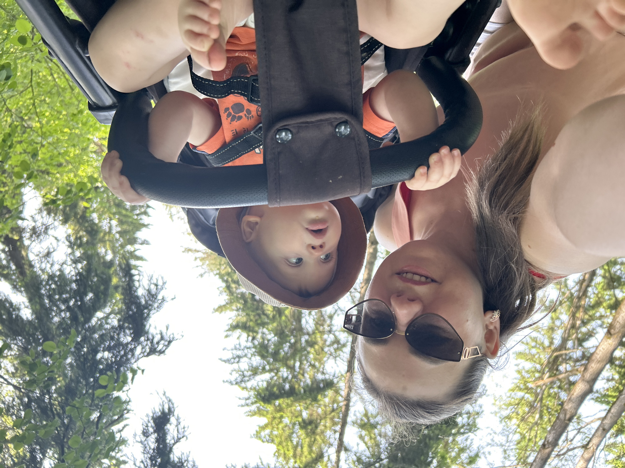 The image is an upside-down selfie taken from a low angle. A woman with sunglasses and shoulder-length hair is on the right side of the frame. To her left, a baby wearing a hat and orange shirt is suspended in a black stroller. The baby's bare feet are visible at the top of the frame. The background is a mix of trees and sky, suggesting an outdoor setting, possibly in the city of Chaikovsky, Russia.