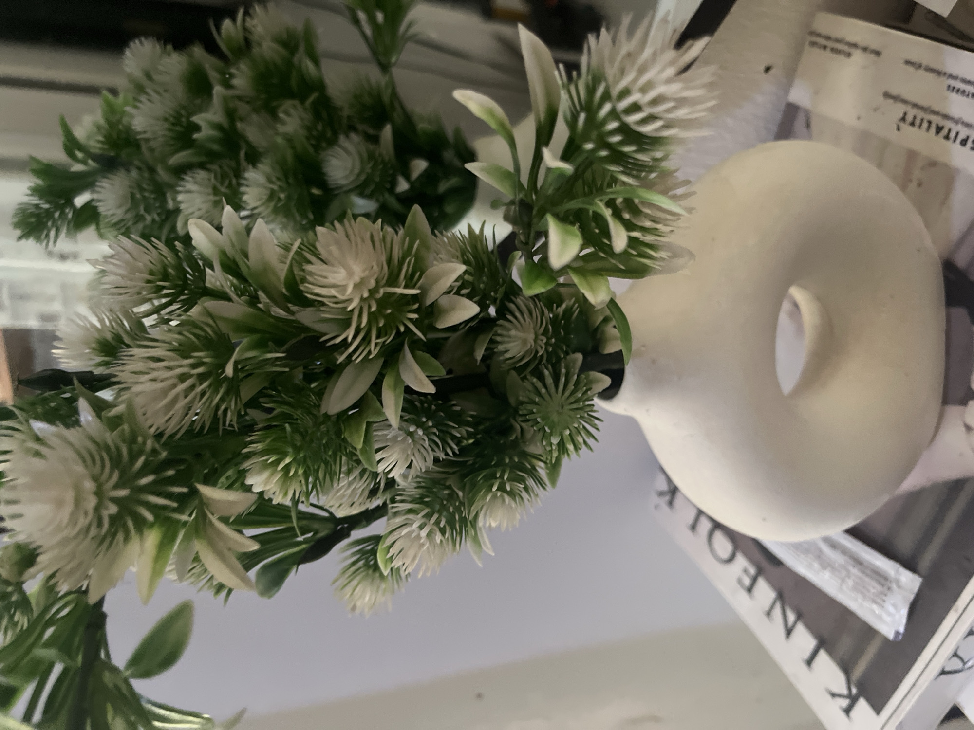 A close-up, eye-level shot captures a faux floral arrangement in a matte white, torus-shaped vase. The plant features numerous small, white, spiky flower-like elements interspersed with green leaves and stems. The vase sits on top of a magazine, the spine of which is visible and reads "KINFOLK" in large, black lettering. The background is a softly blurred, light gray wall, suggesting an indoor setting. No people are present in the image. The overall impression is one of minimalist decor, likely in a home or office environment. No specific location in Abuja, Nigeria, can be determined from the visual information.