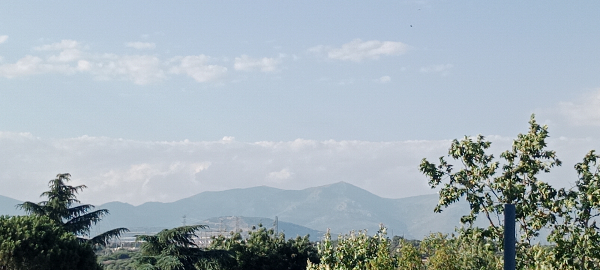 This is a panoramic, eye-level, medium-wide shot of a landscape with mountains in the background and trees in the foreground. The sky is a light blue with scattered white clouds. The mountains are a soft blue-green, suggesting they are in the distance. In the foreground, several green trees are visible, with one particularly prominent tree on the right side of the image. The overall impression is one of a natural, slightly hazy, outdoor environment, likely in a rural or semi-rural setting. The image does not contain any people, actions, or interactions. Given the context of Galapagar, Spain, this likely depicts the natural scenery surrounding the town, possibly a view of the Sierra de Guadarrama mountain range.