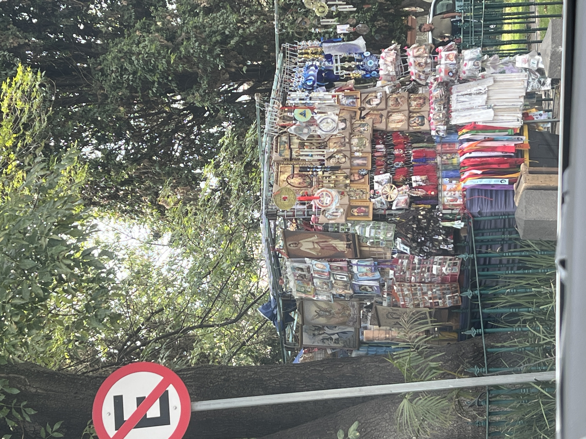 The image shows a vendor's stall selling a variety of religious items and souvenirs in Mexico City. The stall is packed with small framed pictures, keychains, and other trinkets. In the background, there are lush green trees and a "No Parking" sign. A metal fence is visible to the right.