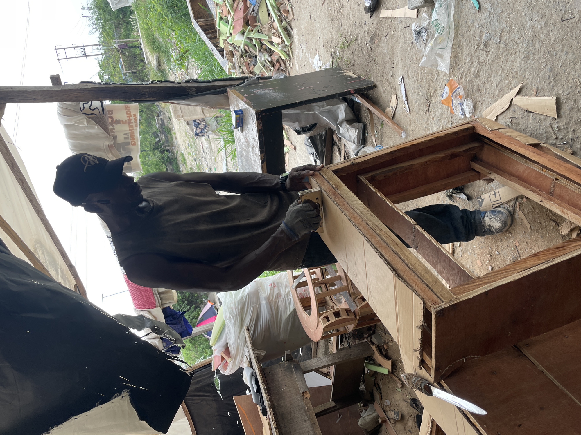In Ojomu, Nigeria, a man wearing a baseball cap, t-shirt, pants, and work gloves is using a staple gun to attach a piece of wood to a wooden box. The scene appears to be in a cluttered, makeshift workshop or living space constructed from wood and tarps. Discarded materials, including wood scraps, cardboard, and plastic, are scattered around the man and the project. A section of greenery can be seen in the background, suggesting an outdoor or semi-outdoor location.