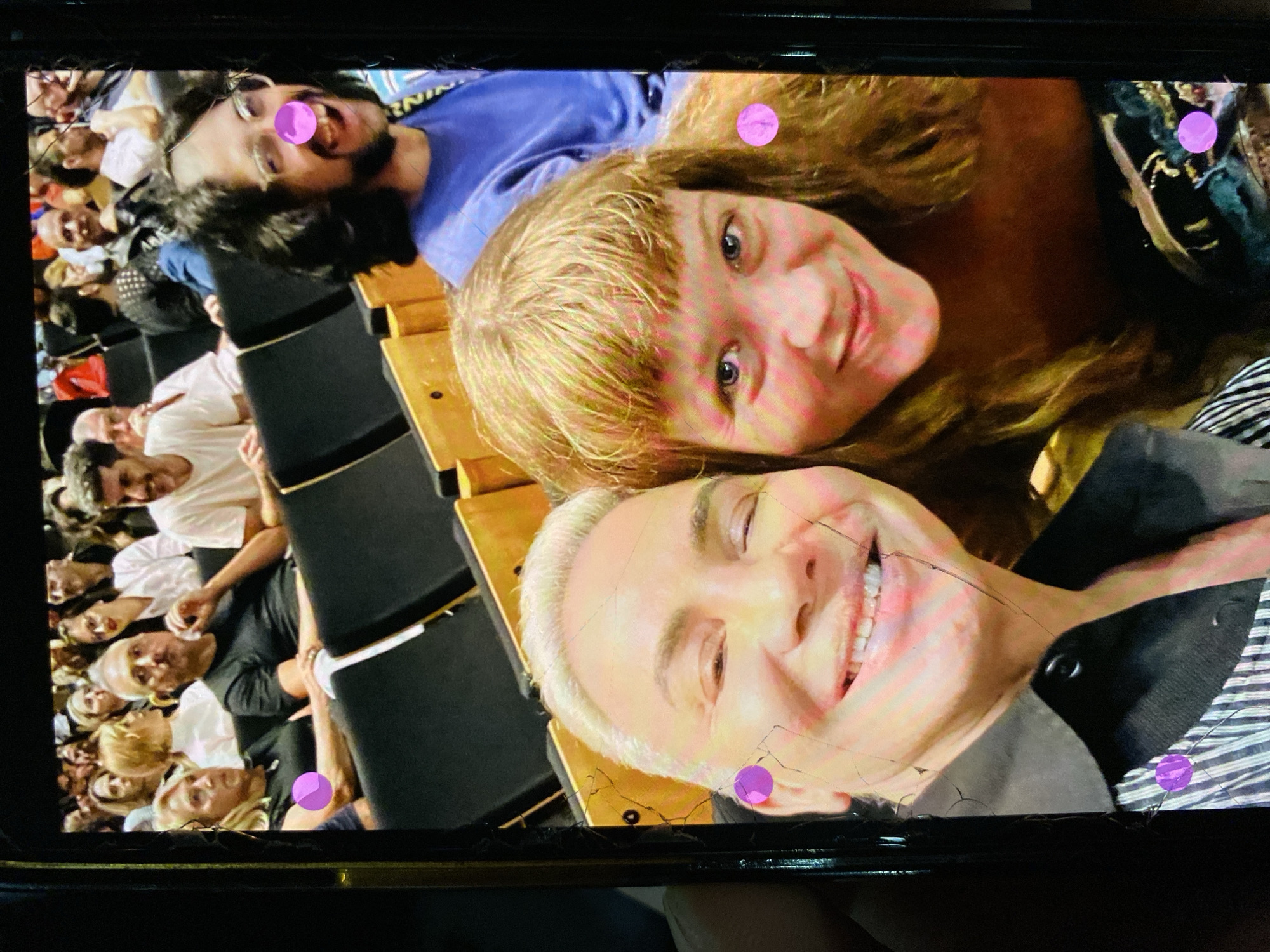 The image displays a photograph taken of a digital screen, showing three smiling individuals in the foreground, with an audience gathered in an auditorium in the background. The entire image is rotated approximately 90 degrees counter-clockwise.
In the foreground, from top to bottom (as per the image's orientation), are three people posing for what appears to be a selfie. The uppermost person is a man with dark, curly hair and a beard, wearing glasses and a blue shirt, smiling broadly with his mouth open. A bright pink circular dot is superimposed over his face. Below him is a woman with light brown/blonde hair, bangs, and blue eyes, smiling gently. Another pink dot is near her forehead. The lowermost foreground individual is a man with short, platinum blonde hair, sporting a wide, cheerful smile that shows many teeth. He wears a striped shirt under a dark garment, possibly a jacket or vest. Two pink dots are visible, one on his ear and another near his shoulder.
The background shows numerous out-of-focus people seated in rows within an auditorium or theater, suggesting they are an audience attending an event, performance, or lecture. Dark seats with lighter wooden accents are visible. The image of the screen itself exhibits several cracks, particularly noticeable across the face of the lowermost man and the dark border around the main photograph.
The subjects in the foreground appear happy and engaged. There are no direct visual cues within the image to specify the time of day, but the setting is clearly indoors. The pink circular dots appear to be digital annotations or filters on the screen image. Some indistinct, blurry text can be seen on a blue background in the upper left corner (as oriented), possibly part of a sign or logo, showing fragmented letters like "RNI". The context provided indicates the location is Buenos Aires, Argentina, though this is not visually confirmed in the image itself.