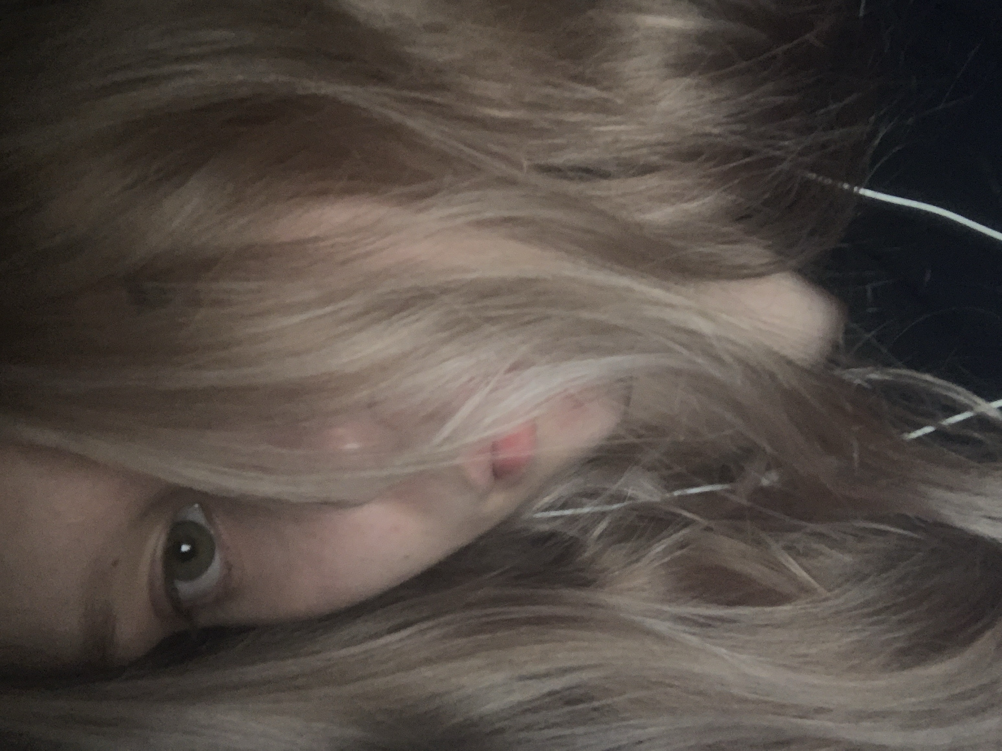The close-up, horizontally oriented image captures a portion of a person's face, predominantly framed and largely obscured by long, wavy, light brown or blonde hair. The subject appears to be lying down, with their head resting, and looks out towards the viewer. The left eye, from the viewer's perspective, is clearly visible, revealing a green or hazel iris with a prominent pupil and a small reflection of light. Parts of the nose and the upper lip, displaying a natural pinkish-red color, are also discernible through the strands of hair.
The extensive hair covers much of the face and acts as a soft, textured element across the frame. In the upper right quadrant, against a dark, undifferentiated background, a thin white wire or cable is visible, emerging from the hair, suggesting the presence of electronic devices such as headphones or a charger. The lighting is dim and soft, consistent with an indoor setting, possibly during the night or in a subdued room.
The expression conveyed by the visible eye is subtle, hinting at a state of observation, contemplation, or perhaps weariness. No specific activity is taking place beyond the implied state of rest or quiet observation. There are no identifiable location cues within the image itself to indicate Saint Petersburg, Russia, and no visible text. The image evokes an intimate and somewhat mysterious atmosphere.