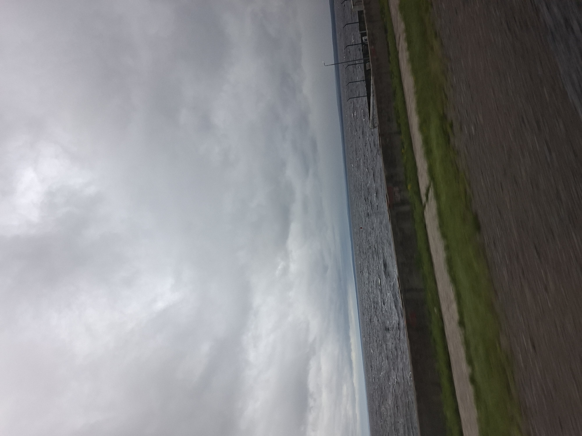 The image depicts a body of water with a cloudy sky overhead. The water appears choppy, suggesting it might be windy or that a boat has recently passed through. Along the right side of the image, there is a paved area with a strip of grass. A railing or fence runs along the edge of this paved area, parallel to the water. In the distance, the water stretches out to meet the horizon, which is obscured by the overcast sky. The overall impression is one of a somewhat gloomy or overcast day, possibly in a park or along a waterfront promenade. There is no visible text in the image. The crops provide closer views of the water, the cloudy sky, and the paved area with grass.