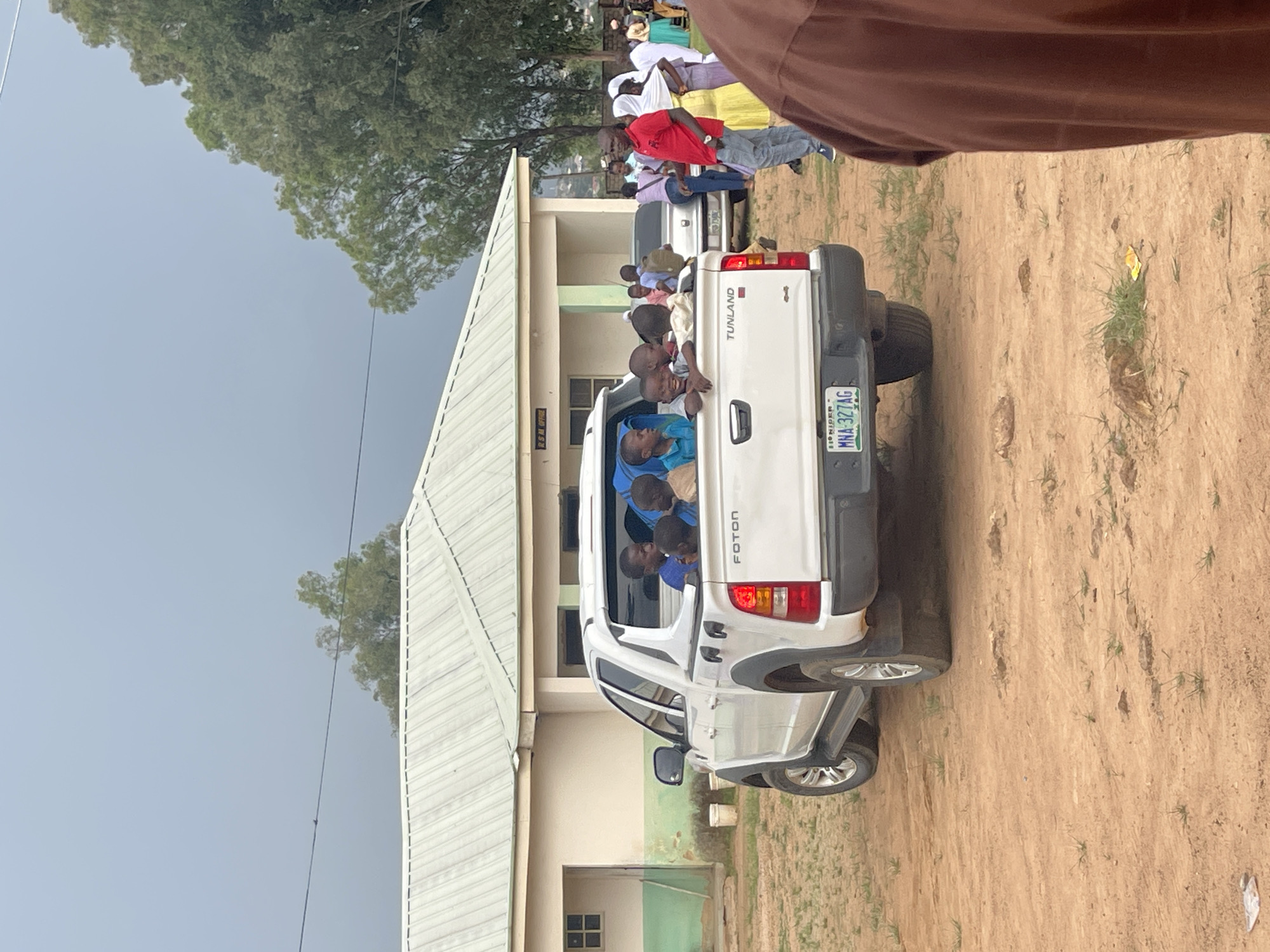 A white Foton pickup truck, with the license plate MNA 327AG, is parked on a dirt road in front of a light-colored, single-story building with a corrugated metal roof. Several young children are visible in the bed of the truck, looking towards the camera. The building appears to be an office, with a sign above the entrance reading "RSM OFFICE". There are other people in the background, some standing and some near other vehicles. The environment is dry and dusty, with sparse vegetation. The sky is overcast. The scene is likely in Unguwar Saya, Nigeria, given the context provided.