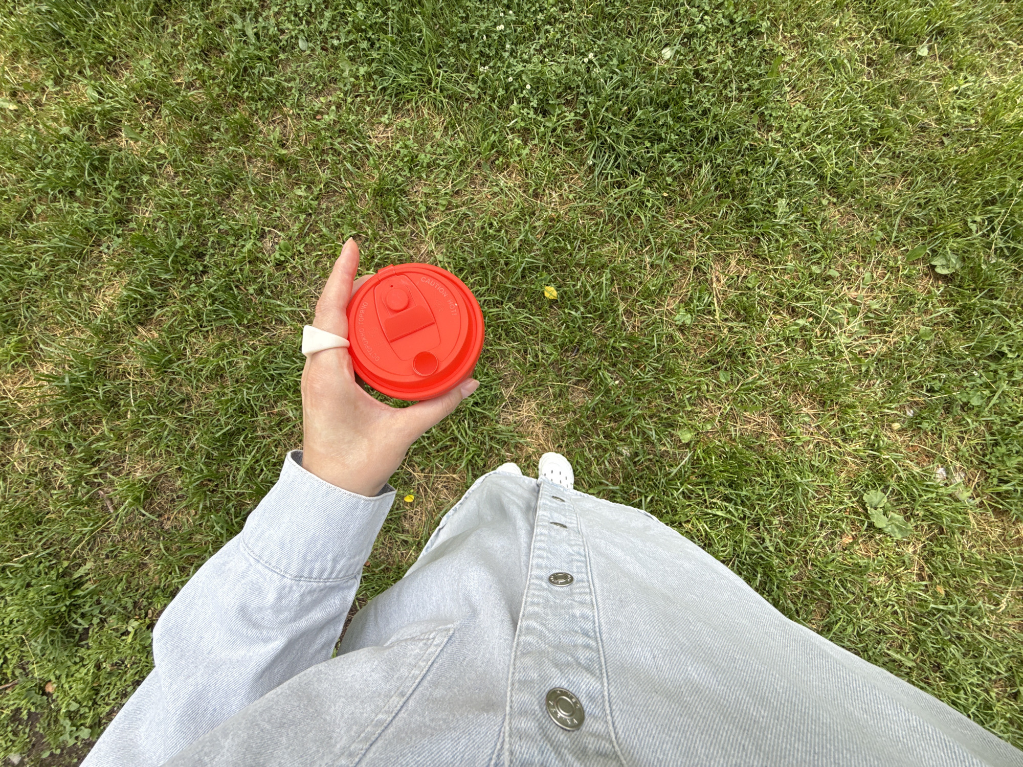 In Izhevsk, Russia, a person wearing a light blue denim jacket and pants holds a bright red travel mug with a white ring on their index finger. The mug has text that appears to be in Russian, reading "ОСТОРОЖНО ГОРЯЧО" which translates to "CAUTION HOT". The scene is set outdoors on a grassy area, with the grass appearing green and somewhat dry in patches. The perspective is from above, looking down at the person's hand and the ground.