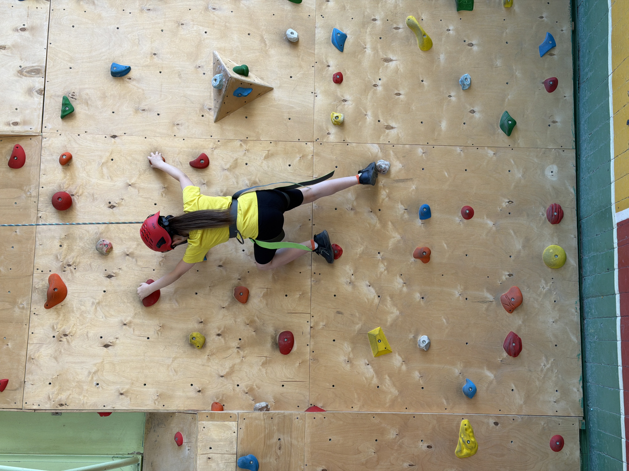 An overhead shot captures a young person actively climbing a wooden rock wall in an indoor setting in Izhevsk, Russia. The climber, wearing a bright yellow t-shirt, black shorts, a red helmet, and a black safety harness with a green strap attached to a blue and white rope, is suspended horizontally against the wall.
The climbing wall is constructed from light-colored plywood panels, densely covered with a variety of multi-colored climbing holds in different shapes and sizes, including red, orange, yellow, green, blue, and gray. A prominent three-dimensional triangular wooden feature, also adorned with holds, is visible in the upper central part of the wall. The climber is in a dynamic pose, with their right hand gripping a red hold and their right foot placed on a gray hold, while their left arm and leg are extended for balance.
To the right of the climbing wall, a section of a painted wall with alternating green, white, and red stripes is visible, suggesting the recreational or athletic nature of the indoor facility. The overall lighting appears consistent with an indoor environment. There is no visible text in the image.