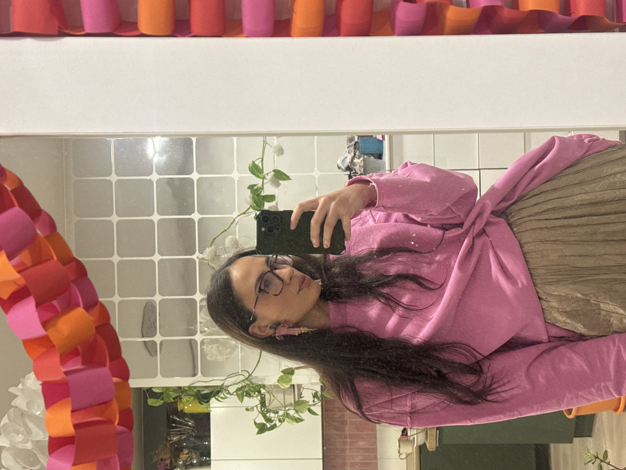 The image depicts a mirror selfie taken by a young woman with long, dark hair and glasses. She is wearing a casual, long-sleeved pink top and a pleated, khaki or olive-green skirt. Her right hand is visible holding a dark-colored smartphone, which she is using to capture the photo. She has distinctive pink and gold tassel earrings. Her expression is neutral and focused on the camera.
The background, visible through the mirror's reflection, features a white tiled wall with square tiles, some of which appear glossy. A green plant with small white flowers is reflected behind her, adding a touch of nature to the indoor setting. Above the mirror's reflection, a plain white shelf or surface spans the width of the frame. To the left and top edges, a vibrant decorative garland crafted from interconnected red, pink, and orange paper loops is visible, along with a large white paper flower partially seen on the left. The scene is located in Krasnoyarsk, Russia. There is no visible text in the image.