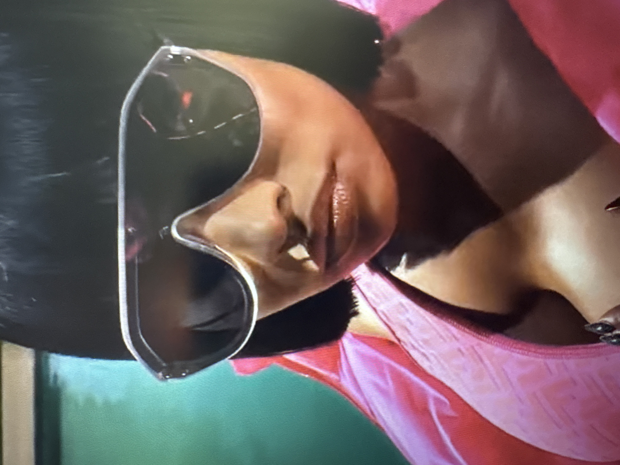 The image shows a close-up of a woman wearing large, silver-rimmed sunglasses and a pink jacket. Her dark hair is cut in a bob. The background is a blurred green wall. The woman's face is angled slightly, and her lips are parted. The image appears to be taken indoors, possibly in a studio or a room with artificial lighting. There is no indication of Sofia, Bulgaria, or any specific city location within the image itself.