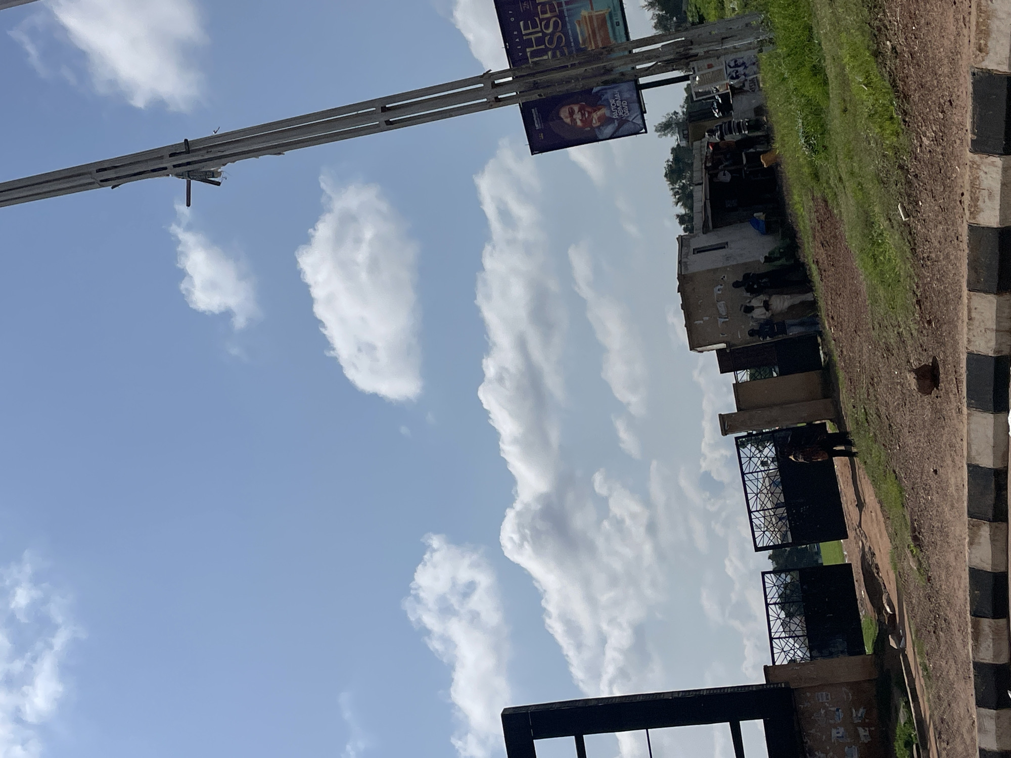 The image depicts a street scene in Jos, Nigeria, featuring a large, open metal gate on the left, likely an entrance to a compound or facility. To the right of the gate, a small, makeshift structure with a corrugated metal roof appears to be a shop or vendor stall, with several people gathered around it. A tall, weathered utility pole stands prominently on the far right, with a large billboard attached to it that displays an image of a woman identified as "PASTOR JENAIAH DAVID" and text that reads "THE ESSEN." The ground in front of the gate is unpaved, with red dirt and some scattered rocks. The sky above is blue with scattered white clouds. A road with a black and white striped curb is visible in the foreground.