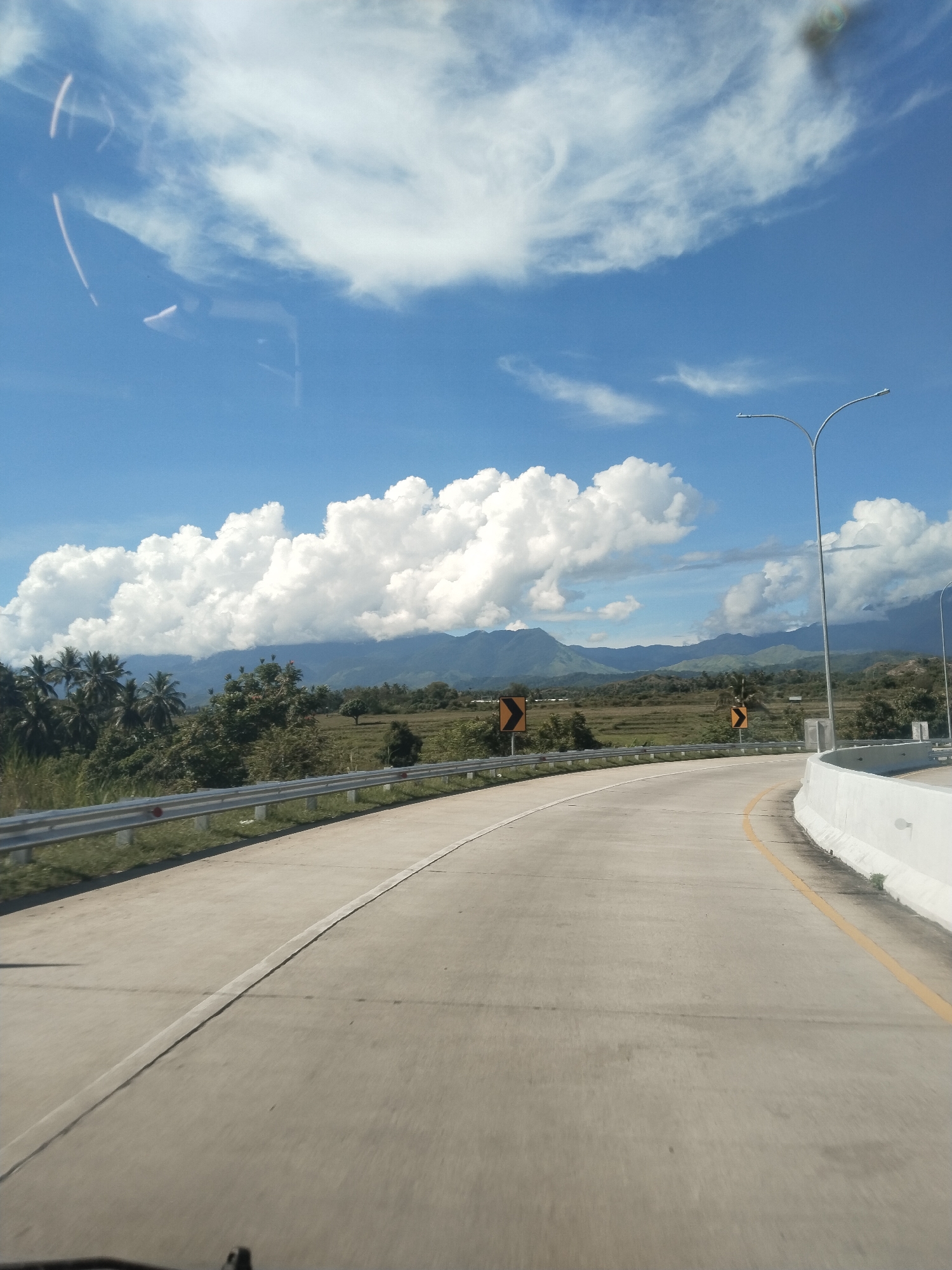 A view from a moving vehicle driving on a concrete road, presumably a highway, near Lampanahtunong, Indonesia. The road curves slightly to the right. A metal guardrail is on the left, and a concrete barrier on the right, with yellow lane markings. Beyond the road, there is a field of low vegetation, possibly rice paddies, with trees and foliage interspersed. A mountain range appears in the background beneath a bright blue sky with puffy white clouds. Streetlights are spaced along the right side of the road. Two yellow road warning signs are visible beyond the guardrail.
