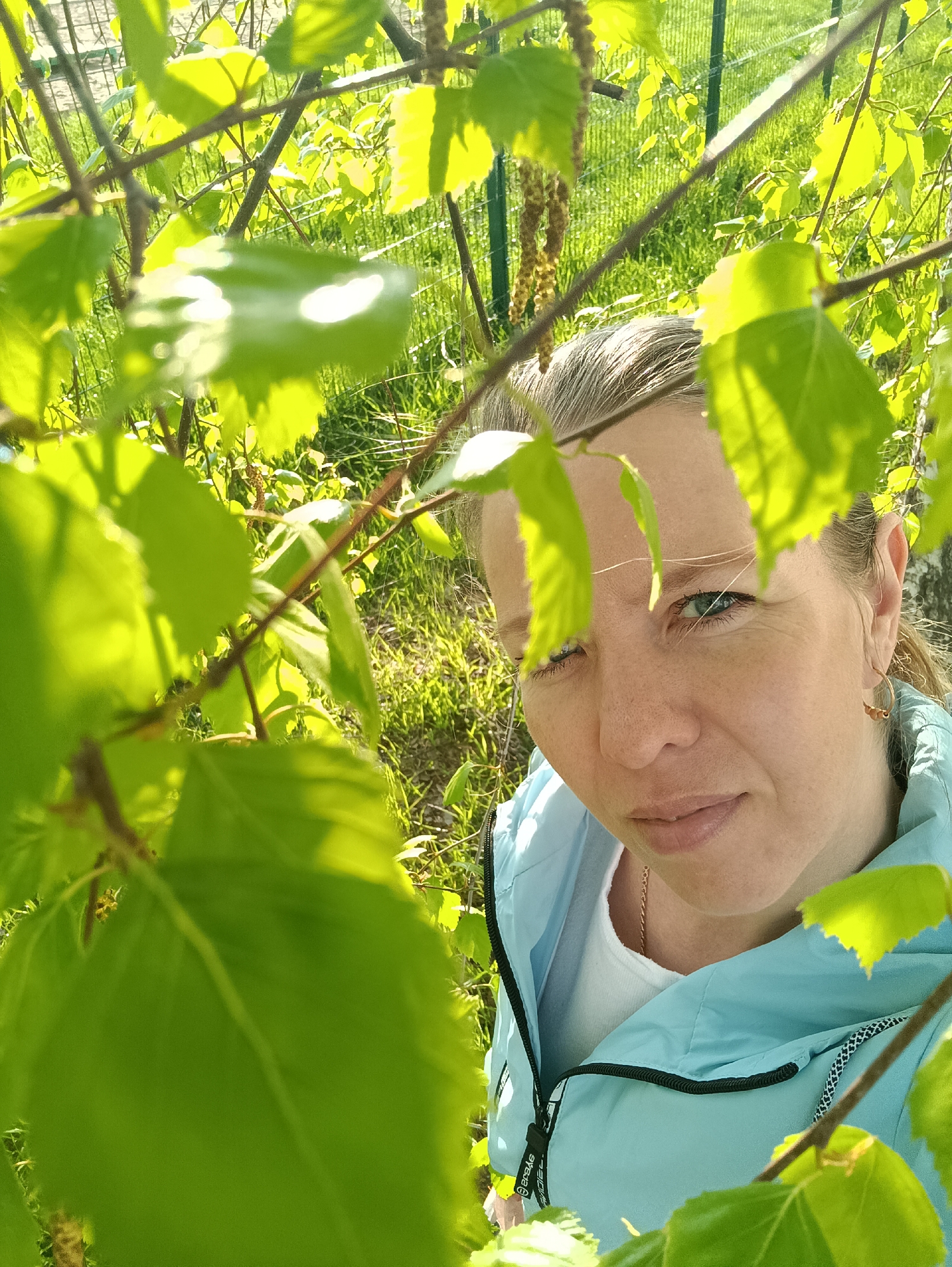 The image shows a woman outdoors in what appears to be a park or garden in Chaikovsky, Russia, based on possible geographical markers. She is positioned close to the camera, partially obscured by tree branches and leaves. She is wearing a light blue jacket over a white shirt and has short, light-colored hair pulled back. Her facial expression is somewhat neutral. The background reveals a grassy area and a fence. The lighting indicates it is daytime, possibly early morning or late afternoon, with strong sunlight highlighting the leaves.