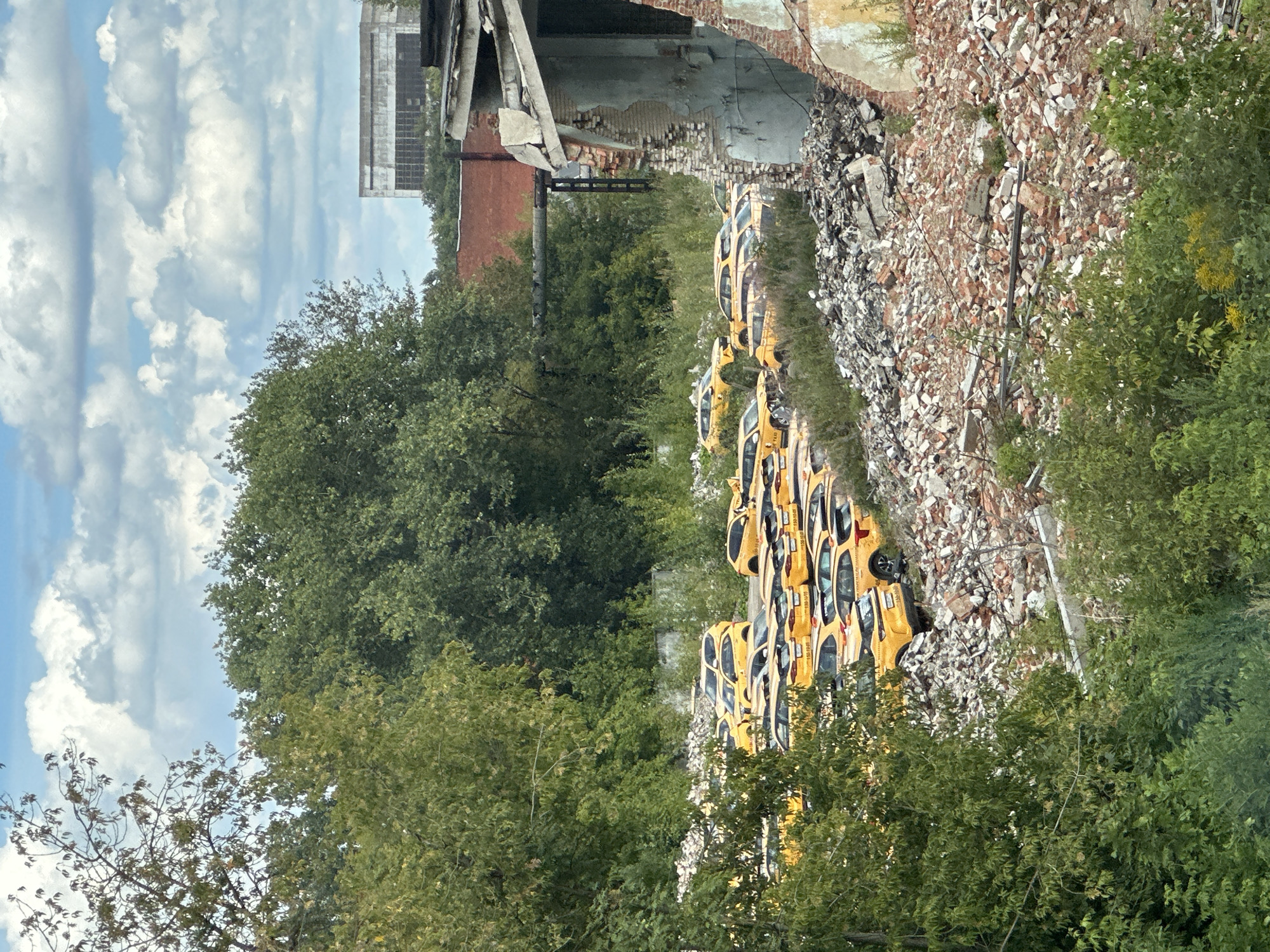 In Podolsk, Russia, an abandoned lot is filled with a large number of yellow taxis. The cars are parked amongst piles of rubble and overgrown vegetation. The scene suggests a derelict area, possibly a former industrial site, with the taxis appearing to be unused or discarded. The background features trees, a cloudy sky, and a decaying building.