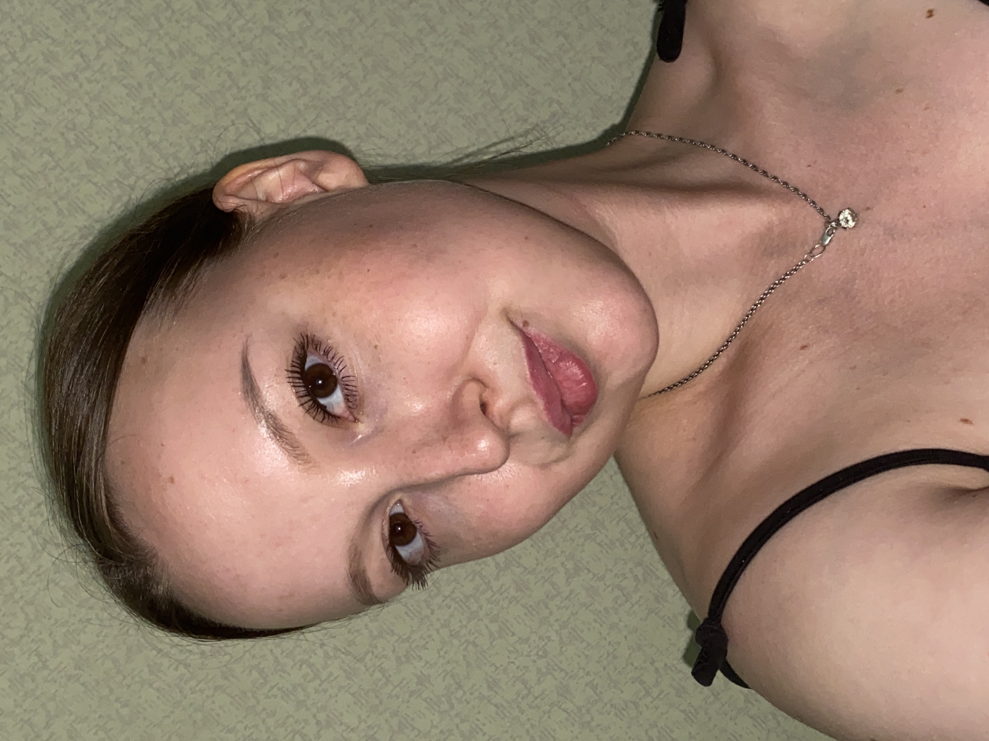 A young woman with brown hair pulled back is looking at the camera. She has brown eyes, light skin with freckles, and pink lipstick. She is wearing a black tank top with thin straps that tie at the shoulders, and a silver necklace with a small pendant. The background is a textured, light green wall. The image is a close-up portrait. There is no indication of the city of Chaikovsky, Russia.