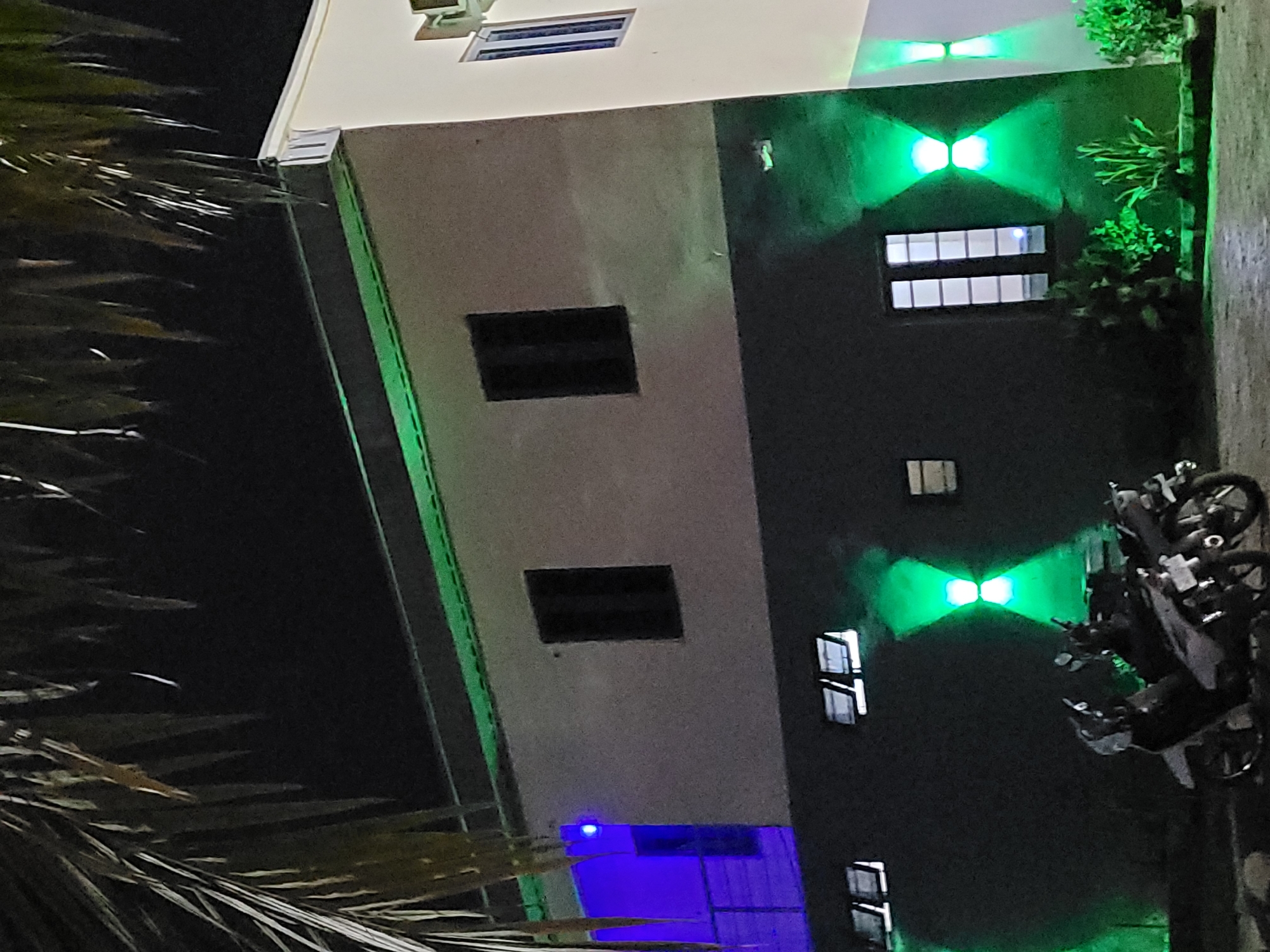 The image depicts a building at night in Akwanga, Nigeria. The building has a mix of beige and dark gray walls, with a green accent stripe running along the edge. Several windows are visible, some lit with green and blue lights, and others dark. Some plants are placed near the windows and on the top edge of the building. A motorcycle is parked to the side of the building. Palm leaves appear in the left foreground, partially obscuring the view.