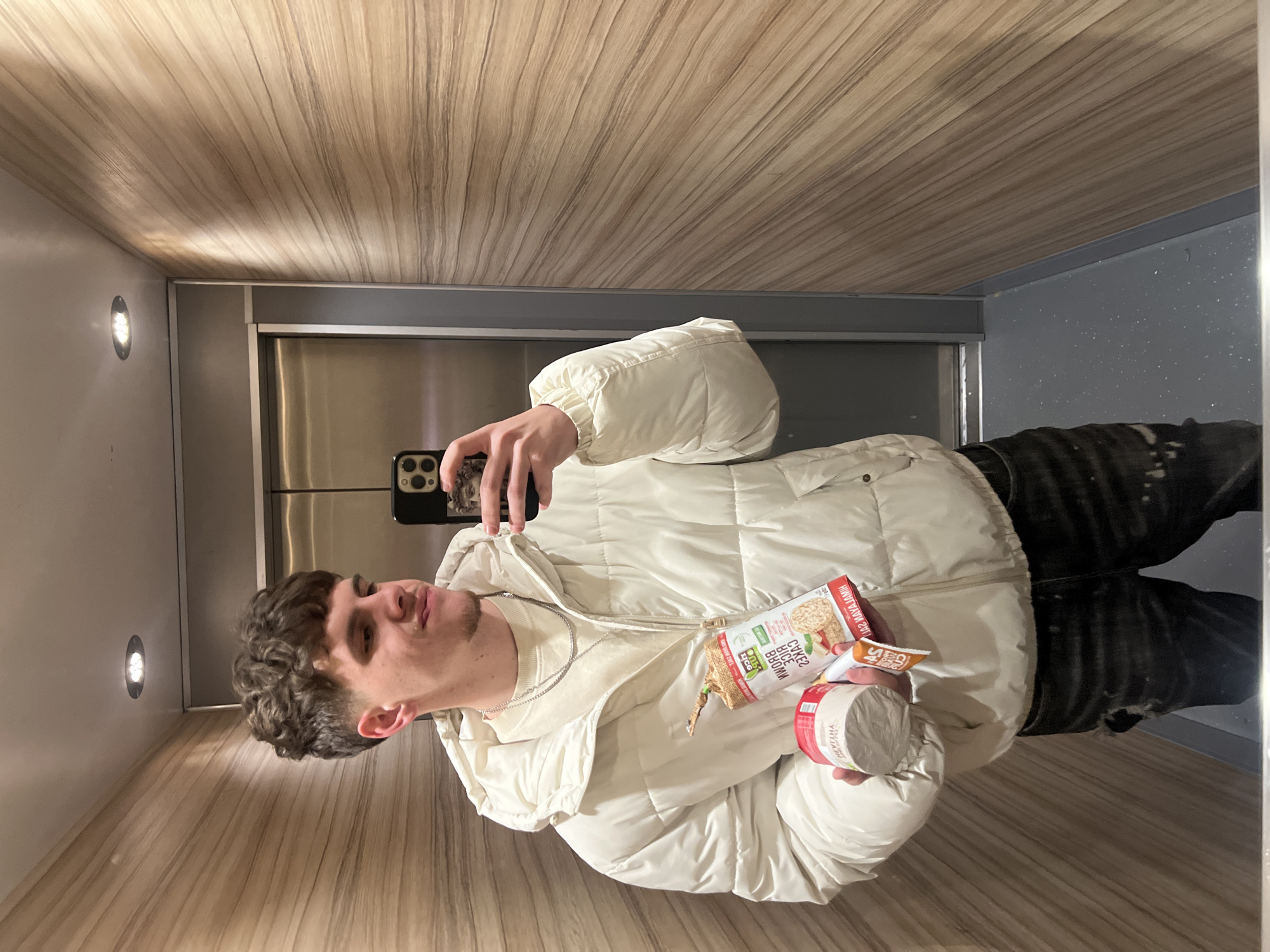 A young adult male is depicted taking a mirror selfie inside an elevator, likely located in Sofia, Bulgaria, as indicated by Bulgarian text on a product package. The male, with short curly brown hair and light facial stubble, wears a white puffer jacket over a light-colored crew-neck sweater and dark distressed jeans with tears and white paint splatters. He holds a black smartphone with a classical bust figurine on its case in his left hand (viewer's right) to capture the image. In his right hand (viewer's left), he holds several items: a white cylindrical container labeled "HEVELTHA," a package of "Dr. Schar BROWN RICE CAKES" with "ОРИЗОВИ ВАФЛИ" (Rice Waffles) in Bulgarian text, and a partially obscured orange pouch with "SPICY CUP" visible. The elevator interior features light wood-paneled walls with horizontal grain, a dark gray speckled floor, and a white ceiling with two recessed circular lights. Metallic elevator doors are visible in the reflection. The subject displays a slight smirk.