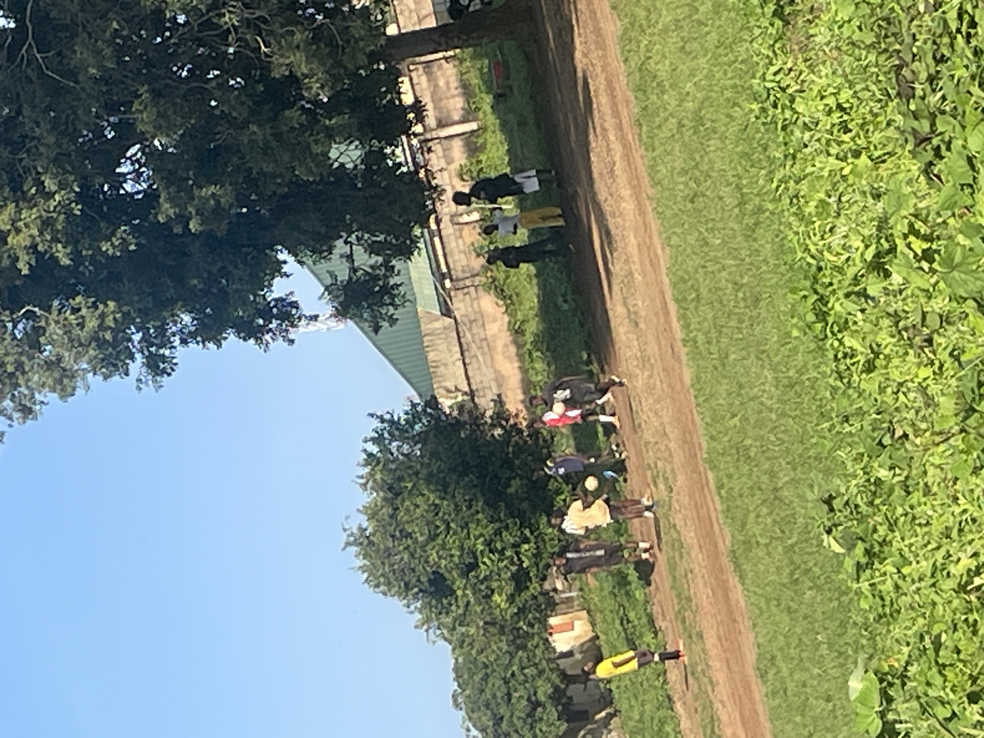 The image captures an outdoor scene with several individuals engaged in a casual soccer game on a dirt field. The environment is characterized by lush green grass in the foreground, a dusty path, and a line of trees and bushes in the background. Beyond the vegetation, a brick wall and a building with a green roof are visible, suggesting an urban or semi-urban setting. The sky is clear and blue. The people are dressed in casual attire, with some wearing soccer jerseys and shorts. One individual in a yellow shirt and black shorts stands out, seemingly observing the game. The overall impression is one of a leisurely activity taking place in a somewhat unkempt, yet vibrant, outdoor space, possibly a local park or a school field in Jos, Nigeria.