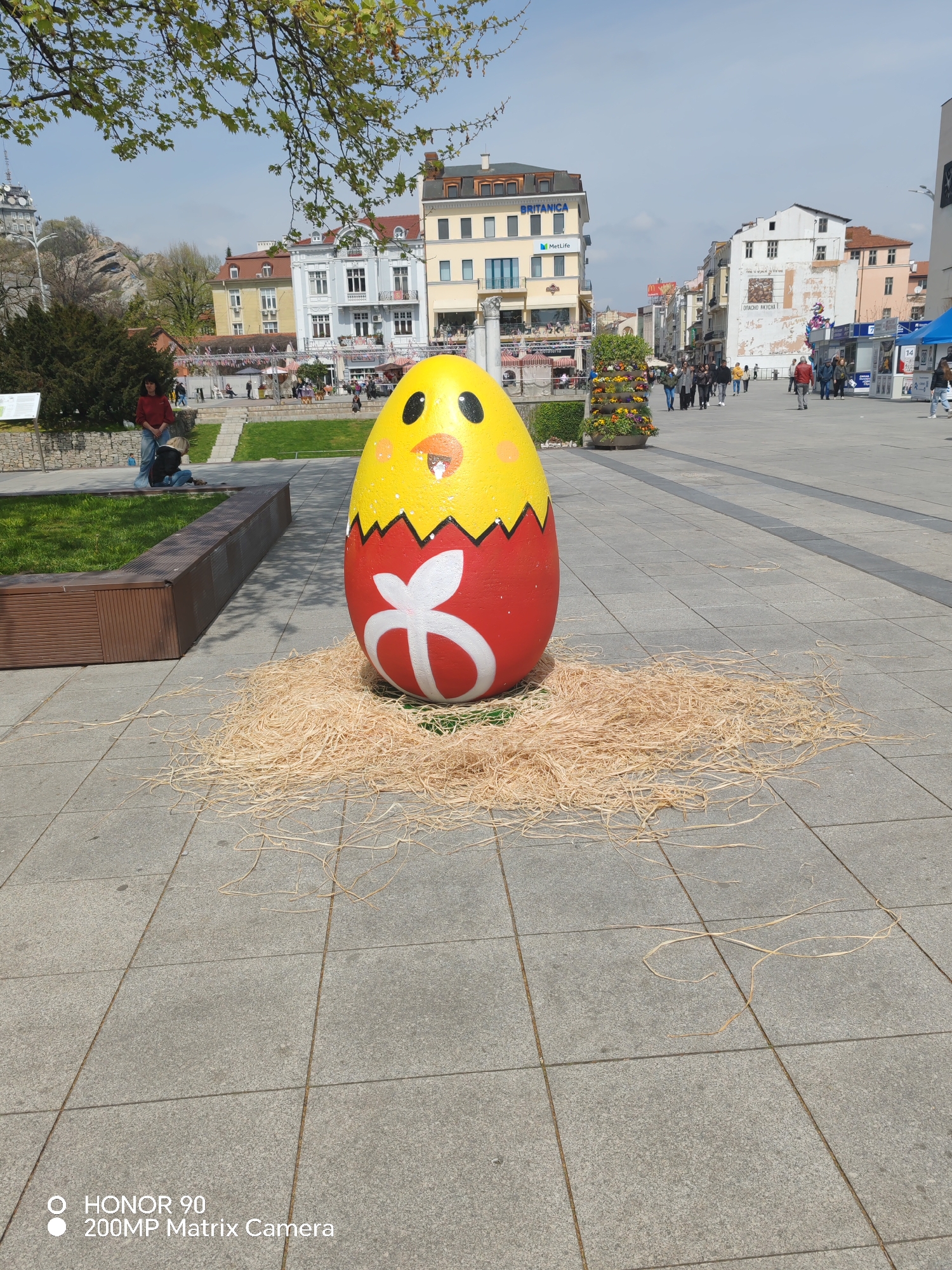 The image depicts a large, decorated Easter egg positioned on a paved public square in Plovdiv, Bulgaria. The egg is painted yellow and red, resembling a chick emerging from its shell, and features a white floral design. It rests on a bed of straw. In the background, there are several buildings with varied architectural styles, a public garden with trees, shrubs, and people walking around. A woman and child are seated near a wooden bench, and other people are visible walking in the distance. There is a visible smartphone watermark indicating the image was taken with an Honor 90 camera.
