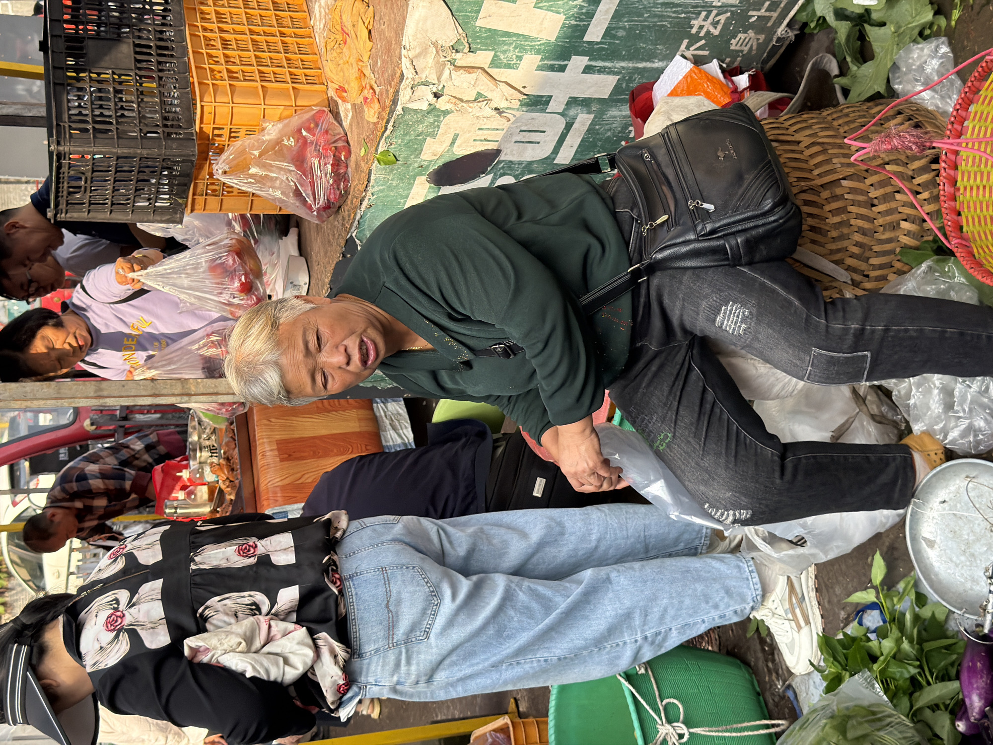 The image captures a street scene in Jiangjin, China, likely a market setting, with a focus on a middle-aged woman seated on a woven basket. She has short grey hair and is wearing a dark green long-sleeved shirt and black jeans. A black leather purse with silver accents is slung across her body. Her mouth is slightly open as if she is speaking.
Behind her, another woman, from the back, is wearing a black jacket with a pattern of faces and roses, and light blue jeans. She is holding a white cloth. Several plastic bags filled with red items, possibly fruit, are visible in the background, along with a red and white plastic crate. Other market stalls and people are blurred in the background, suggesting a busy environment. The ground is littered with plastic bags and some vegetables, including eggplants, are in the foreground. The overall scene depicts a vibrant, bustling market with people engaged in commerce.