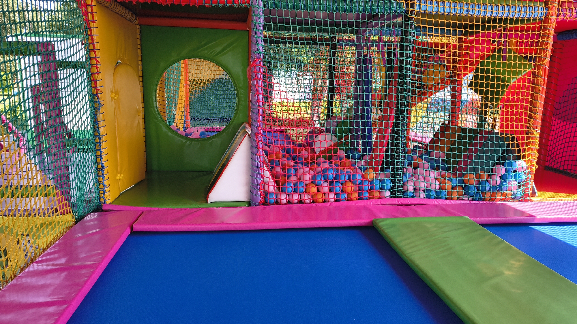 The image depicts the interior of a colorful indoor playground, likely a children's entertainment center. The scene is dominated by bright primary and secondary colors, with soft, padded surfaces and netting creating various play areas. In the foreground, a large blue trampoline surface is visible, bordered by thick pink padding. To the right, a green padded mat lies diagonally across the trampoline. Behind this, a section of the play structure is visible, featuring green, yellow, and pink padded walls, as well as netting enclosures. A prominent feature is a ball pit filled with numerous brightly colored plastic balls, including pink, blue, and orange. A circular opening in a green padded wall allows a glimpse into another area of the playground, also appearing to contain balls. The environment suggests a safe and stimulating space for children's play. The specific location in Mikhaylovka, Russia, is not visually discernible from the image itself but would be an external contextual detail.
