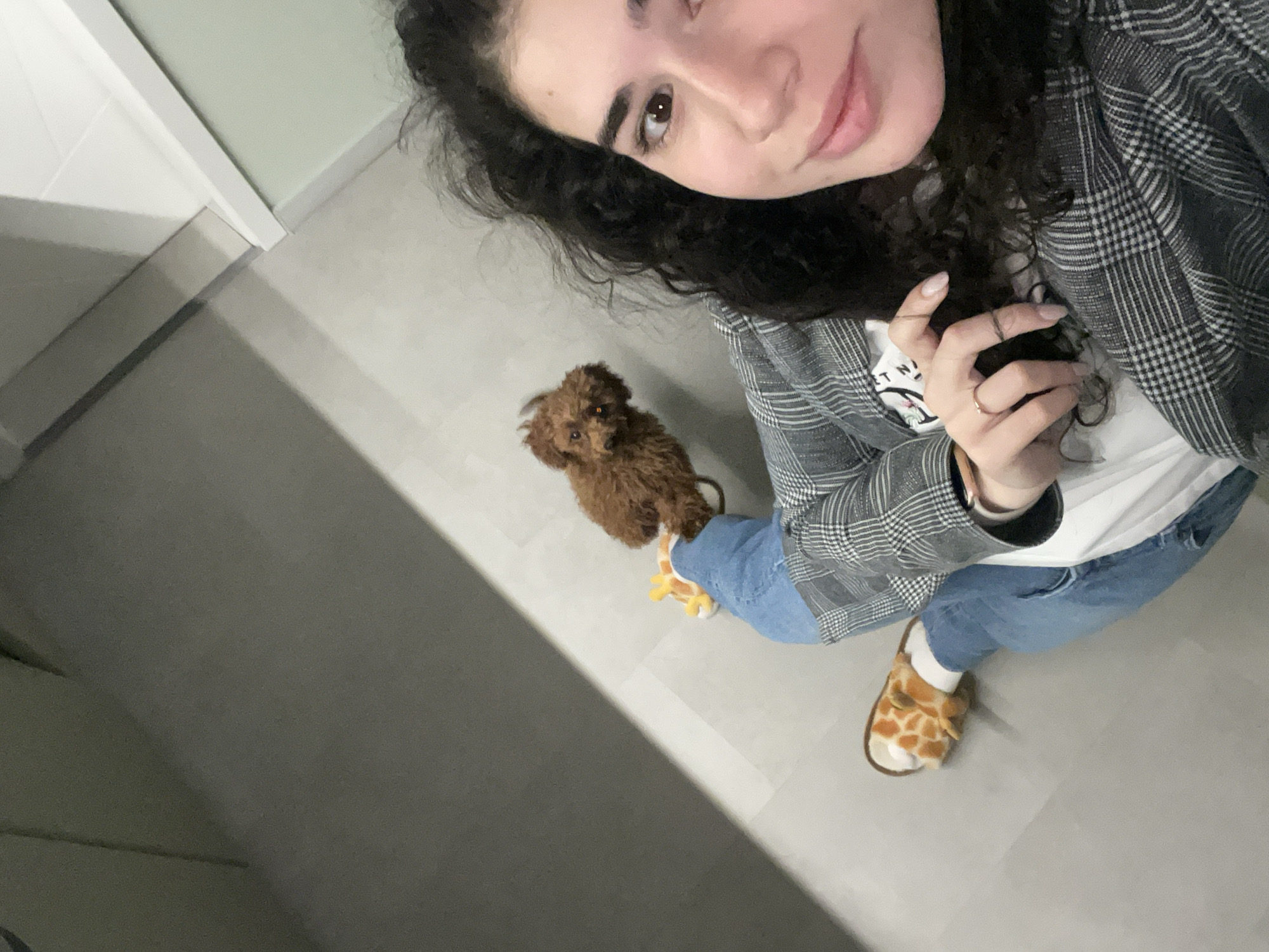 A woman stands indoors with a small brown poodle at her feet. The woman is fair-skinned, with dark curly hair, wearing a gray plaid blazer over a white t-shirt, blue jeans, and giraffe-patterned slippers. She is holding her hand near her face and appears to be taking a selfie. The poodle is small with tightly curled fur and is standing on a stuffed giraffe toy. The setting appears to be a room with light gray flooring. The location in Sofia, Bulgaria is unknown based on the provided image.