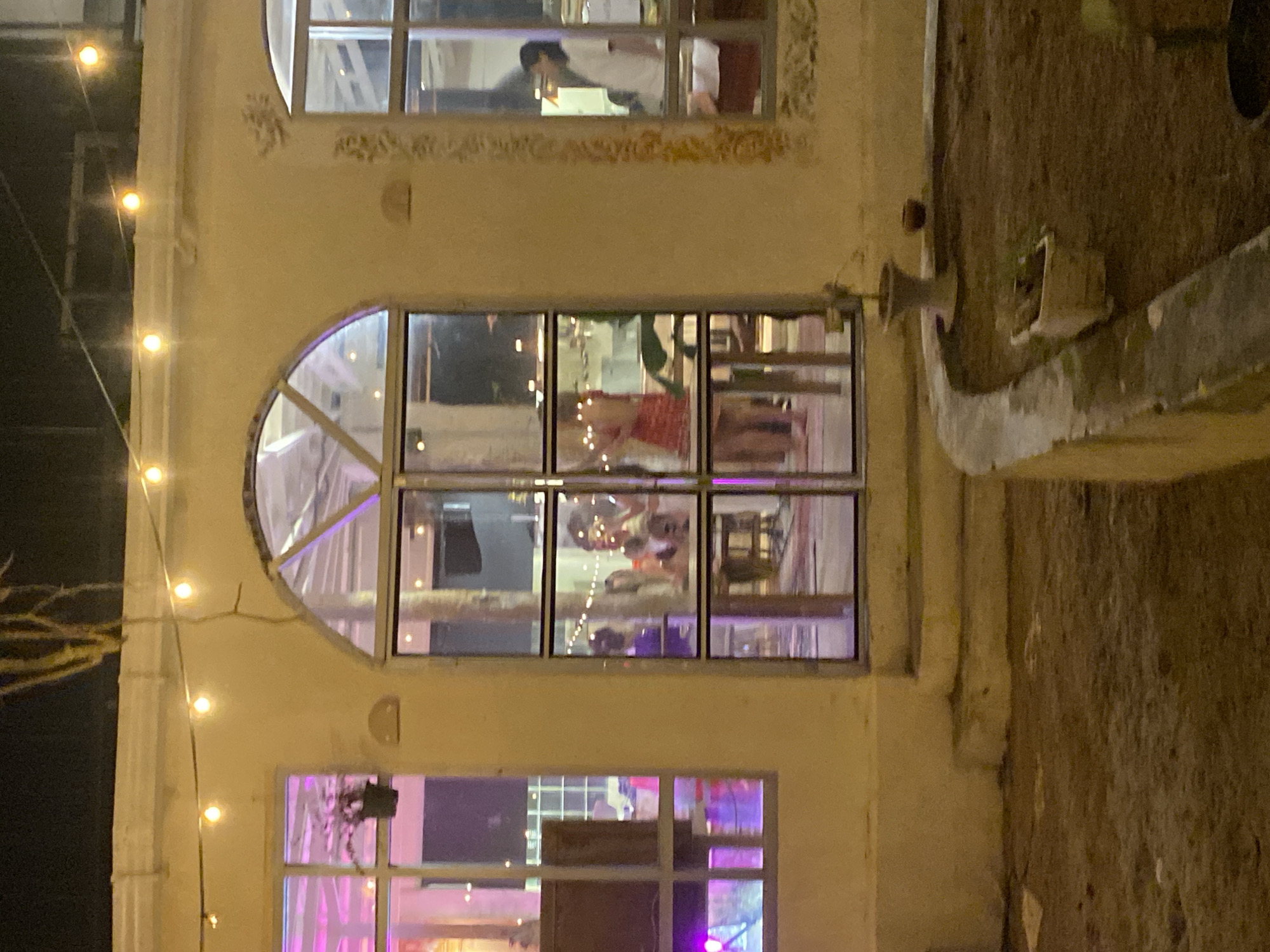 The image shows the exterior of a building at night, illuminated by string lights. The building has multiple windows, some of which reveal interior scenes.
In the top window, a person in a white shirt and dark pants is visible, appearing to be seated at a table with a laptop or book. In the large arched window below, a group of people are gathered. They appear to be socializing, with some individuals looking towards the camera. The interior is lit with warm, ambient light.
The lowest visible window shows a more colorful interior, with purple and pink lighting and what appear to be event decorations. The overall scene suggests a social gathering or event taking place inside the building. The exterior walls of the building are a light yellow color, and a textured, rougher surface is visible to the right of the windows, possibly a wall or a planter. The time of day is clearly night due to the artificial lighting and dark surroundings. There are no visible signs or text in the image.