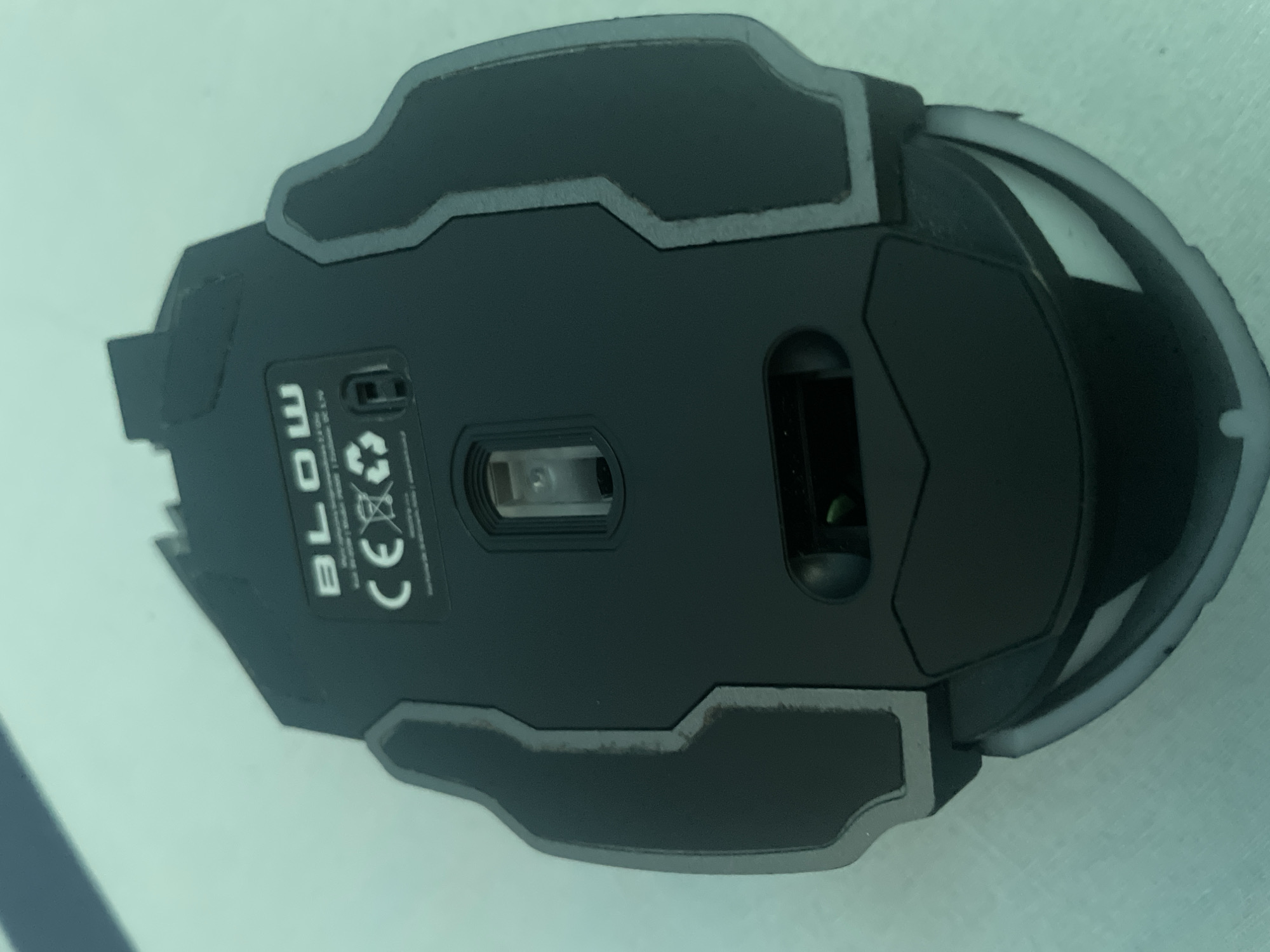 The image shows the underside of a black computer mouse. The brand "BLOW" is visible on the top, along with CE and recycling symbols. A small optical sensor is located in the center of the mouse. There appears to be a compartment for a battery or wireless receiver on the lower part of the mouse, which is slightly ajar, revealing a green wire. The mouse is resting on a light blue, textured surface, possibly a table or desk. The image is taken from a top-down perspective, with the mouse positioned slightly off-center. There is no indication of the city of Kołobrzeg, Poland, or any other location in the image.