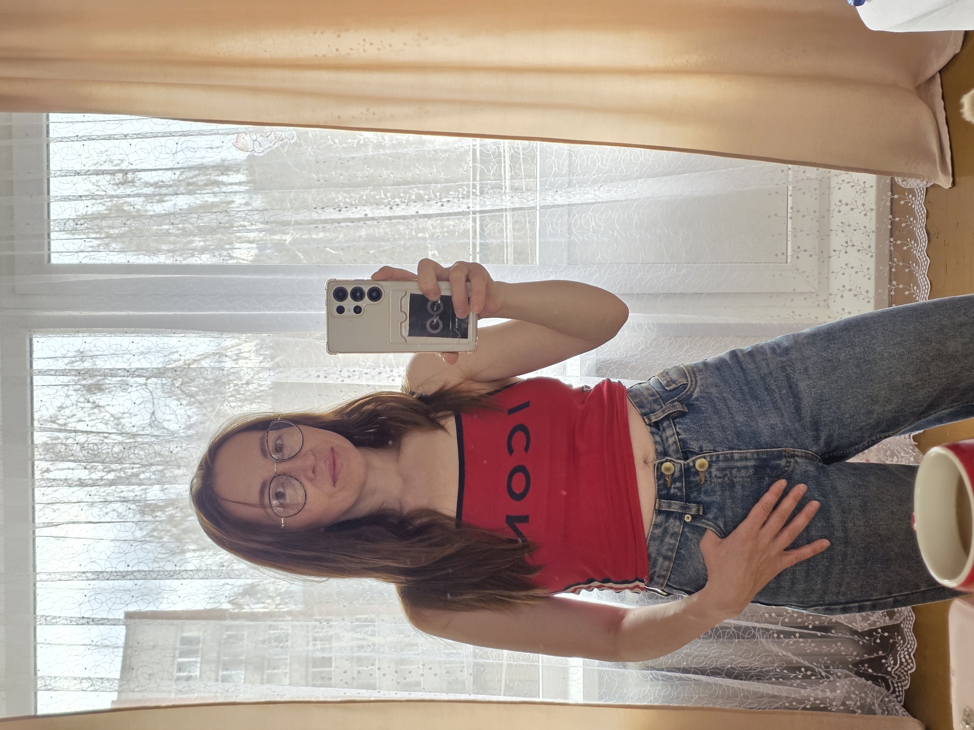 A woman stands indoors, facing a mirror, taking a selfie with a smartphone. She wears glasses, a red tank top with "ICON" printed on it, and blue jeans. Her left hand rests on her hip while her right hand holds the phone. Behind her is a window with sheer, patterned curtains. Outside the window, a building is visible, suggesting an urban environment. The photo was taken in Chaikovsky, Russia.