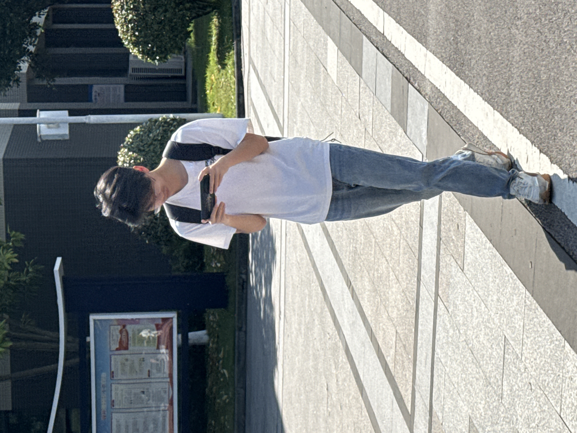 The image depicts a young man standing on a paved outdoor walkway in Jiangjin, China, looking down at a smartphone he holds horizontally with both hands. He is wearing a white short-sleeved t-shirt, blue jeans, and light-colored sneakers, with black backpack straps visible over his shoulders.
The scene takes place on a bright, sunny day, likely late morning or early afternoon, as indicated by the strong shadows cast by the man and nearby objects onto the light-colored, patterned pavement. The walkway is composed of alternating light and dark grey rectangular blocks. Adjacent to the walkway is an asphalt road marked with a distinctive white line.
In the background, there are green bushes and hedges, along with dark-colored buildings. A white-framed notice board or display stand is visible in the lower-left background, featuring several framed documents. Although the text on these documents is illegible, characters (likely Chinese) are discernible, with some red highlights in the upper sections. The overall setting suggests a public space, possibly a campus or urban area in Jiangjin.