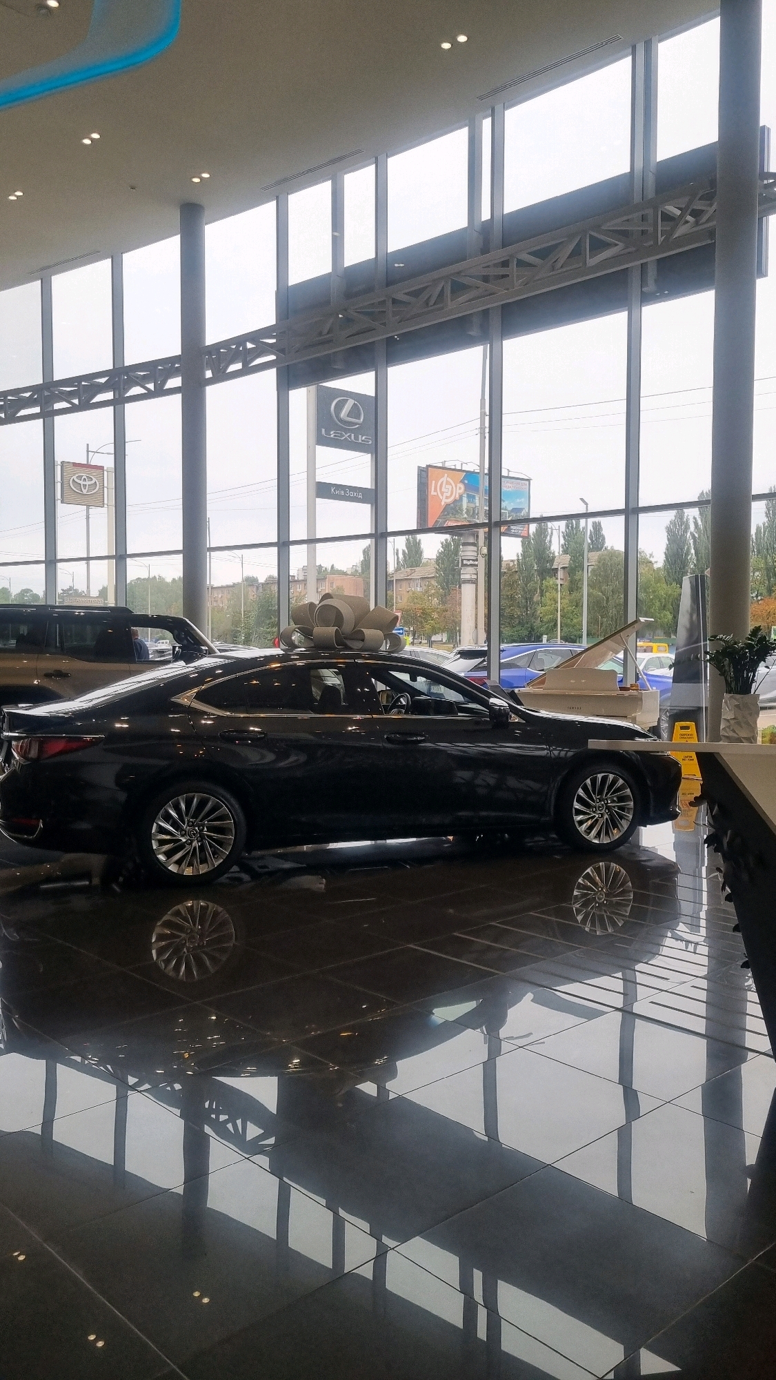 This image depicts the interior of a car dealership, likely a Lexus dealership, given the prominent Lexus logo visible in the background. The main subject is a black Lexus sedan prominently displayed with a large, beige bow on its roof, suggesting it is a new car or a special promotional vehicle. The car is positioned on a glossy, reflective floor, which mirrors the vehicle and the surrounding architecture.
In the background, through large glass windows, an overcast sky and trees are visible, indicating an outdoor environment beyond the dealership. Other vehicles, including what appears to be a white grand piano, a blue car, and a dark SUV, are also present within the dealership, suggesting a wide selection of vehicles or perhaps a special event. Signs for Toyota and what appears to be a local business or highway exit ("Київ Захід" which translates to "Kyiv West") are visible, hinting at the dealership's location. The overall environment suggests a modern and well-lit commercial space. The presence of the Lexus logo and the "Kyiv West" sign strongly suggest the location is in or near Kyiv, Ukraine, with Sofiivska Borshchahivka being a suburb of Kyiv.