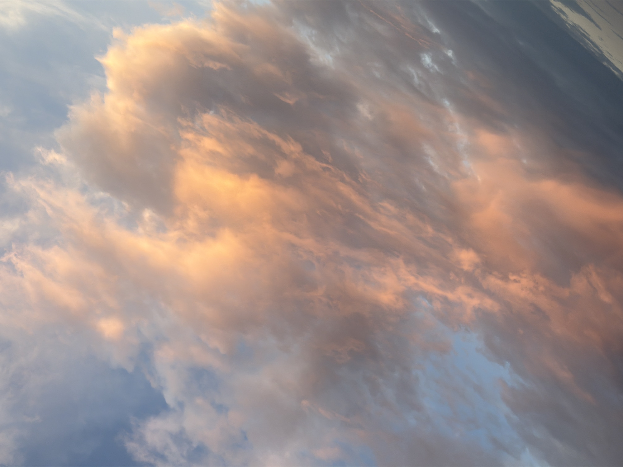 The image shows a sky filled with clouds, likely during sunrise or sunset. The clouds are illuminated with a soft, warm light, ranging from peach to orange hues, while other parts of the sky are a light blue or gray. The composition is mostly clouds, with no visible land or other elements. The image is shot with a tilted angle. There is no visible location in the city of Petrovac na Moru, Montenegro.